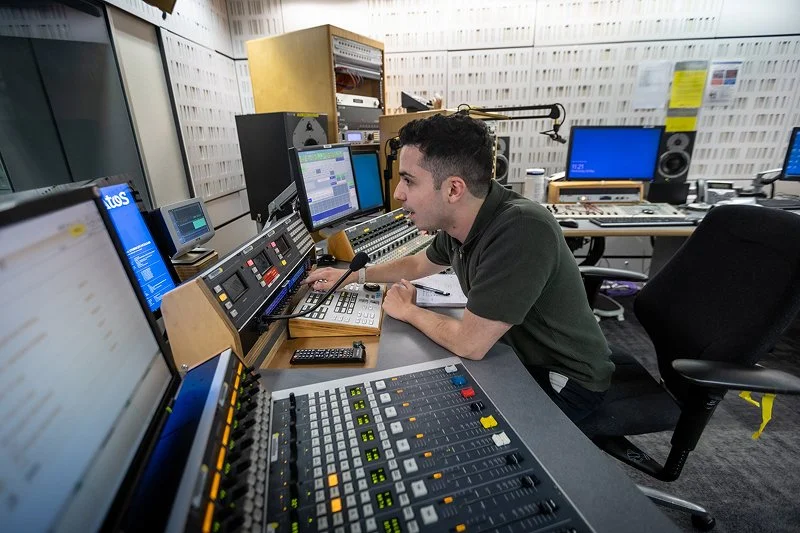 A man working in a radio or recording studio surrounded by sound equipment, screens, and control panels.