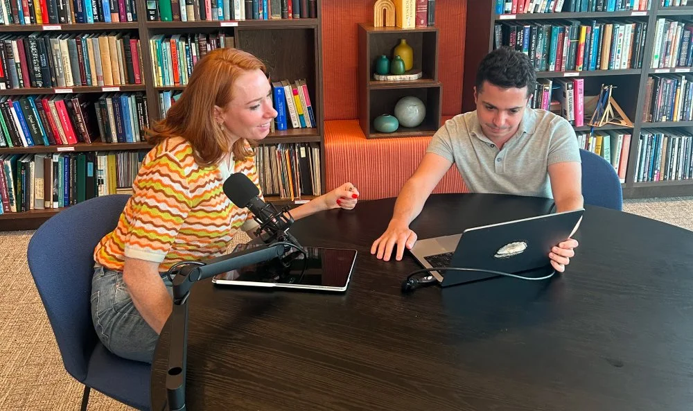 A woman and a man sitting at a black conference table with bookshelves in the background. The woman has red hair, is wearing a multicolored striped shirt, and is speaking into a microphone. The man has curly dark hair, is wearing a gray polo shirt, and is using a laptop.