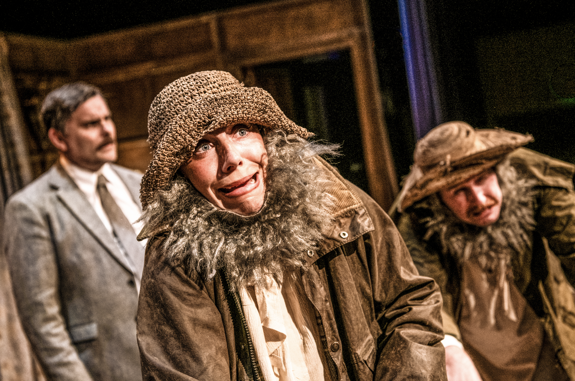 Three actors on stage in a dramatic scene, with the woman in the foreground wearing a brown hat and coat with a fur collar, displaying a distressed or fearful expression. A man in the background wears a similar hat and coat, also with a fur collar, and looks downwards, while another man in the background wears a suit and tie and looks on with a serious expression. The setting appears to be indoors with wooden furniture.