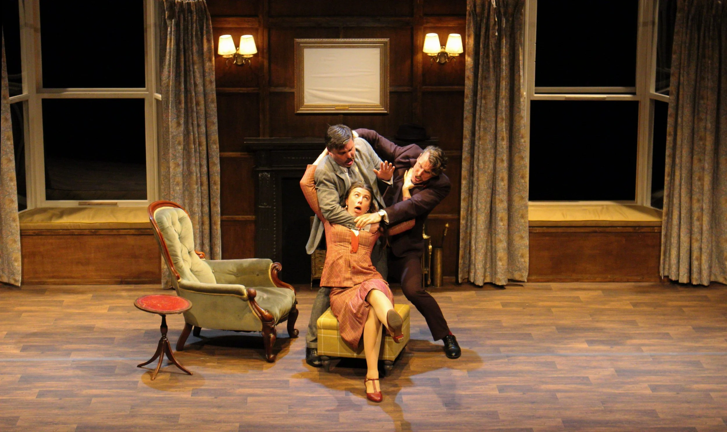 Three actors on a stage performing a dramatic scene. One female seated on a small ottoman, two men grabbing her in an intense confrontation. The stage is decorated like a vintage room with wood-paneled walls, a fireplace, large windows with curtains, and vintage furniture.