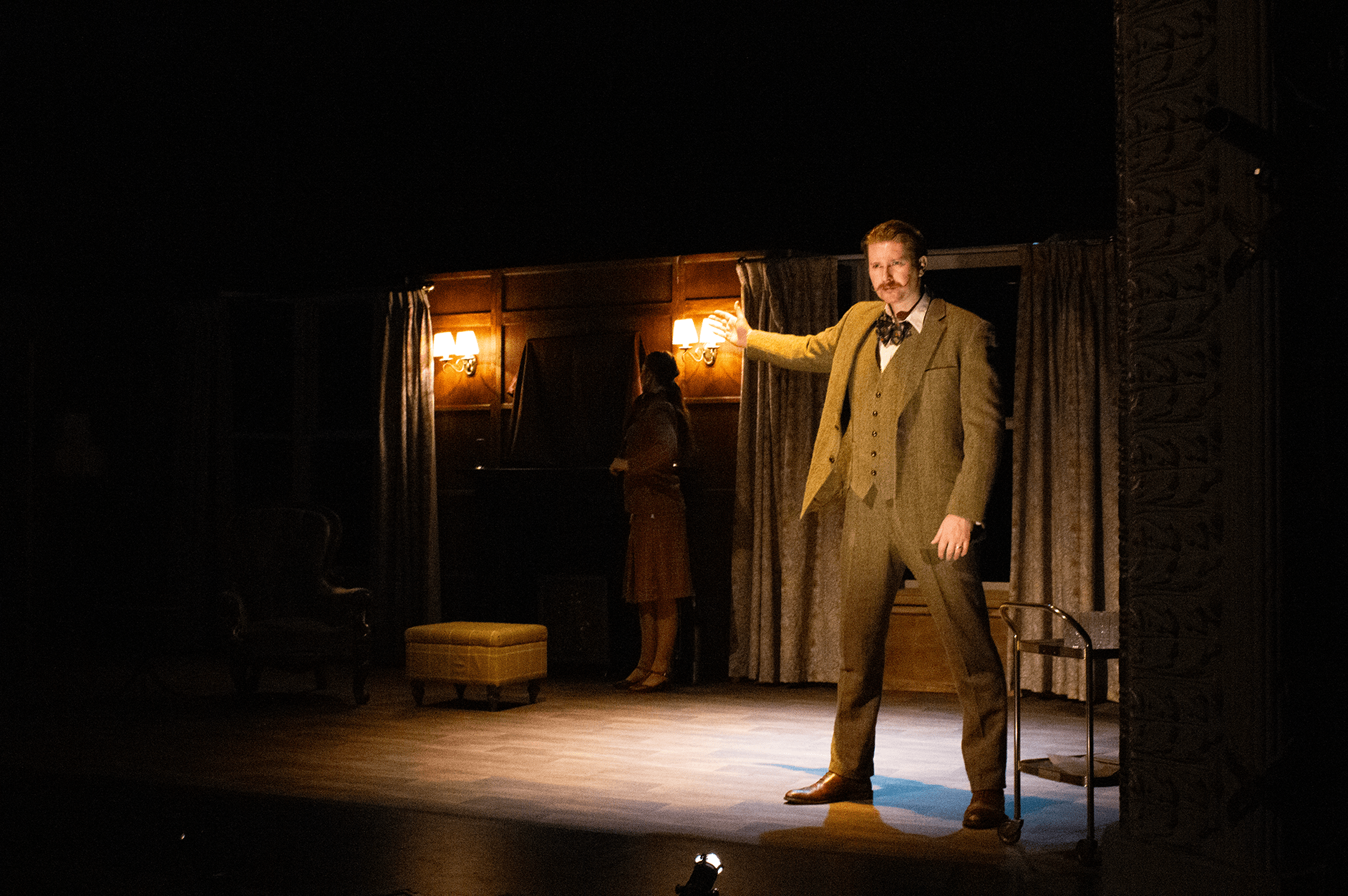 A man in 19th-century attire standing on a stage, gesturing outward, with a woman in period clothing in the background near a piano, in a dimly lit theater or performance space.