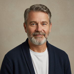 A portrait of a middle-aged man with gray hair and a beard, wearing a navy blue cardigan over a white shirt, standing against a beige background.
