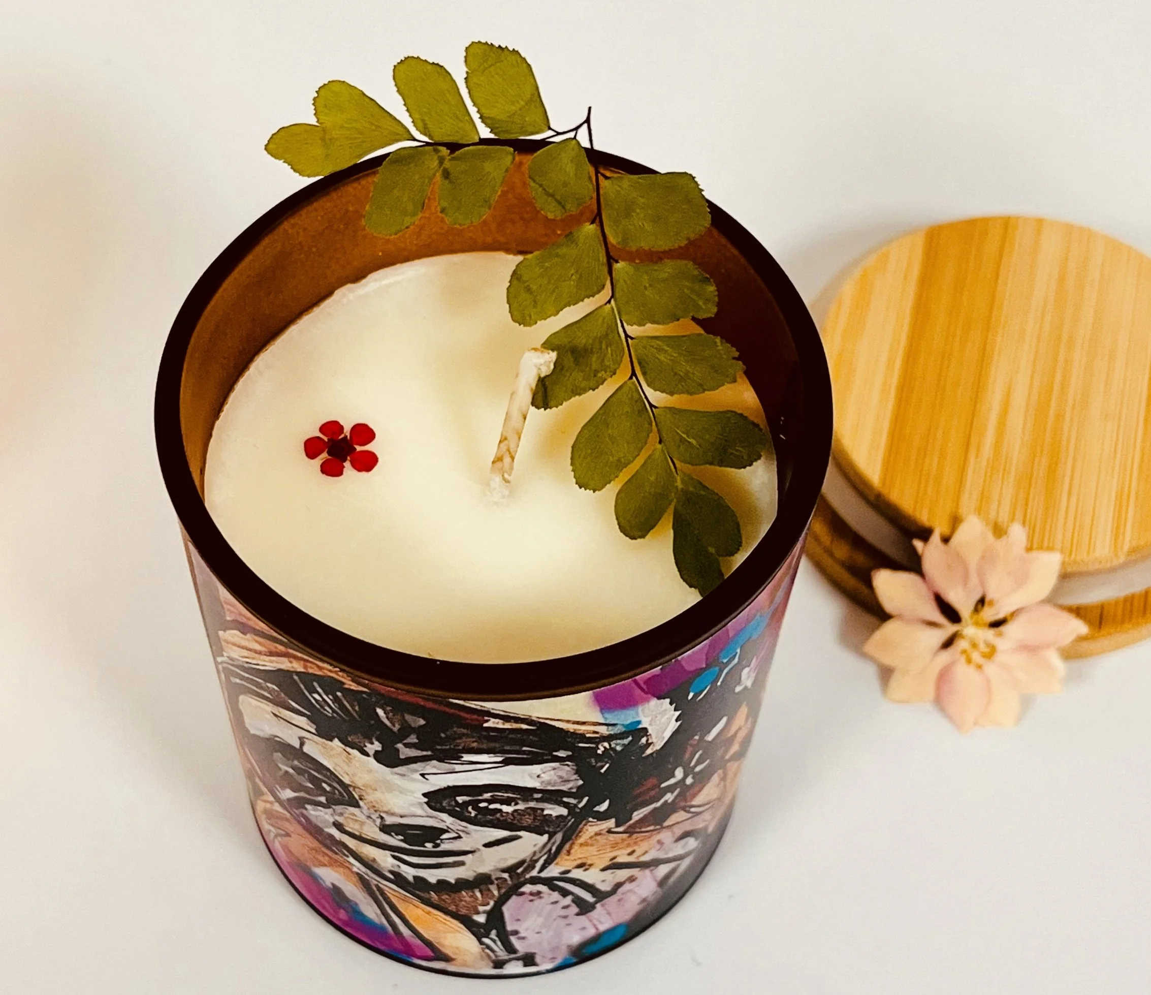 Decorative candle with pink and black abstract art on glass holder, a green fern leaf on top, small red flowers floating in white wax, beside a wooden container with a pink flower decoration.