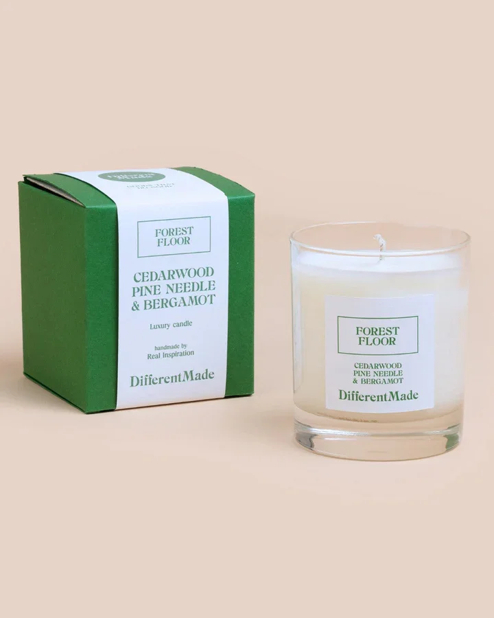 Green and white boxed candle labeled 'Cedarwood Pine Needle & Bergamot' next to a clear glass candle holder with a white candle inside. The labels indicate it is a luxury candle by DifferentMade.