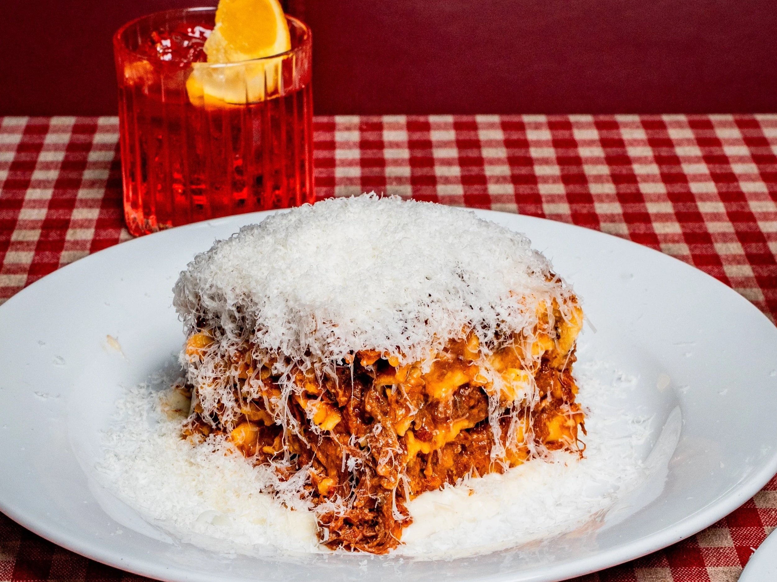 A plate with a serving of lasagna topped with grated cheese, a glass of NEGRONI with an Orange wedge, on a red and white checkered tablecloth.