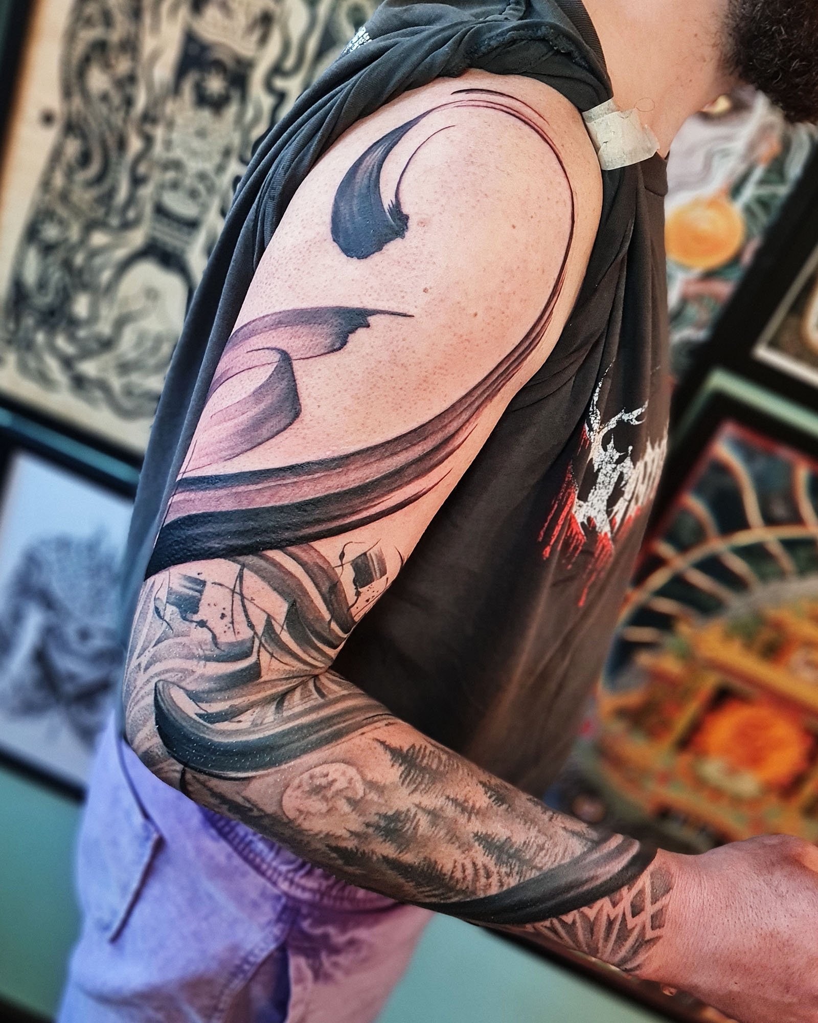 Person with a large, abstract black and grey tattoo sleeve on their left arm, featuring swirling patterns and nature elements like trees and a moon.