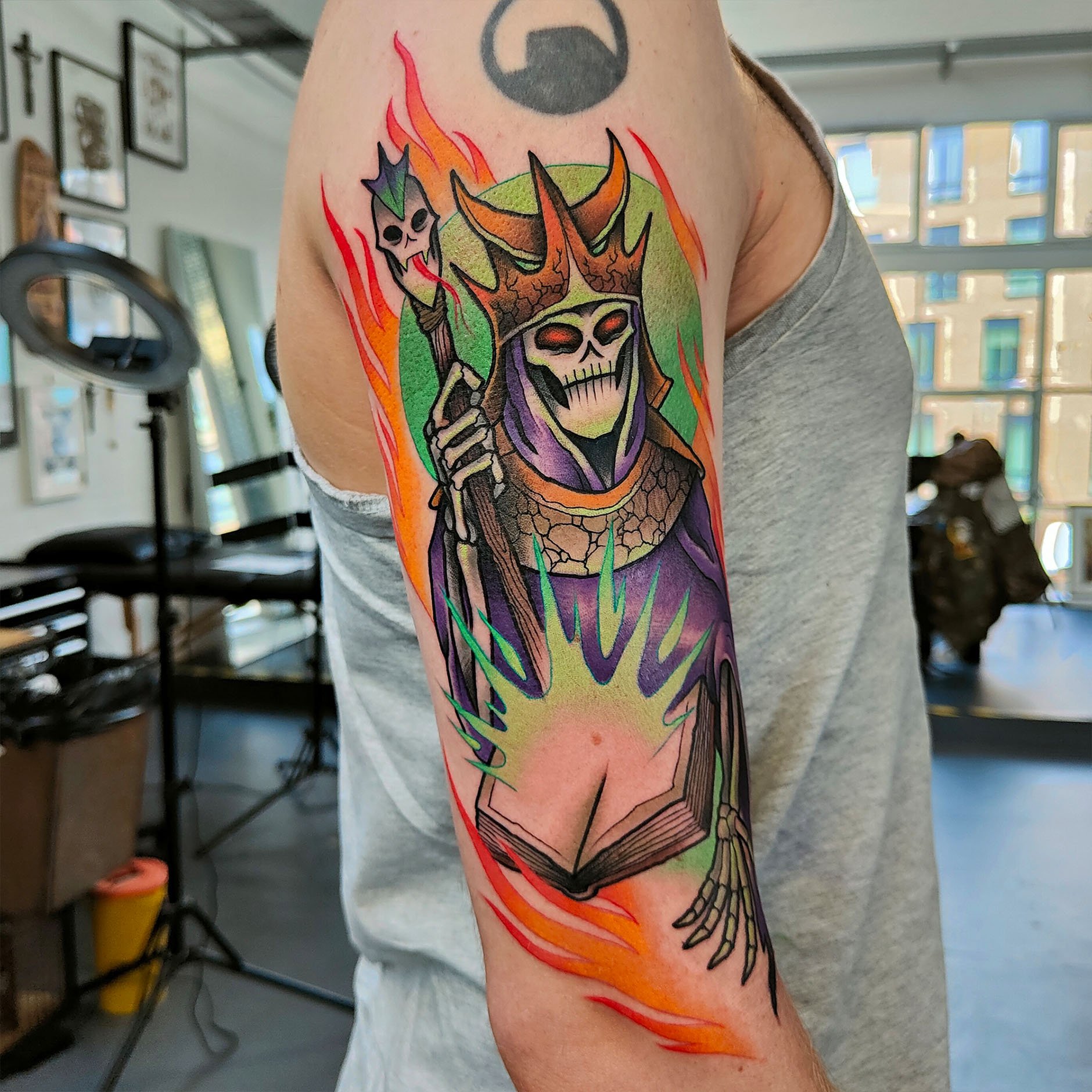 Colorful tattoo on arm featuring a necromancer with a crown, holding a staff with a skull on top, surrounded by flames.