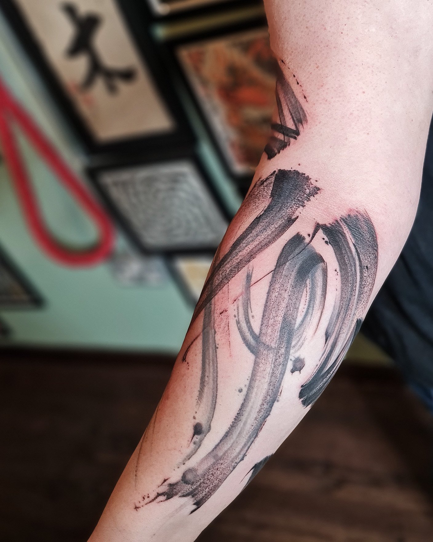 Abstract black ink brushstroke tattoo on inner forearm with art frames in background.