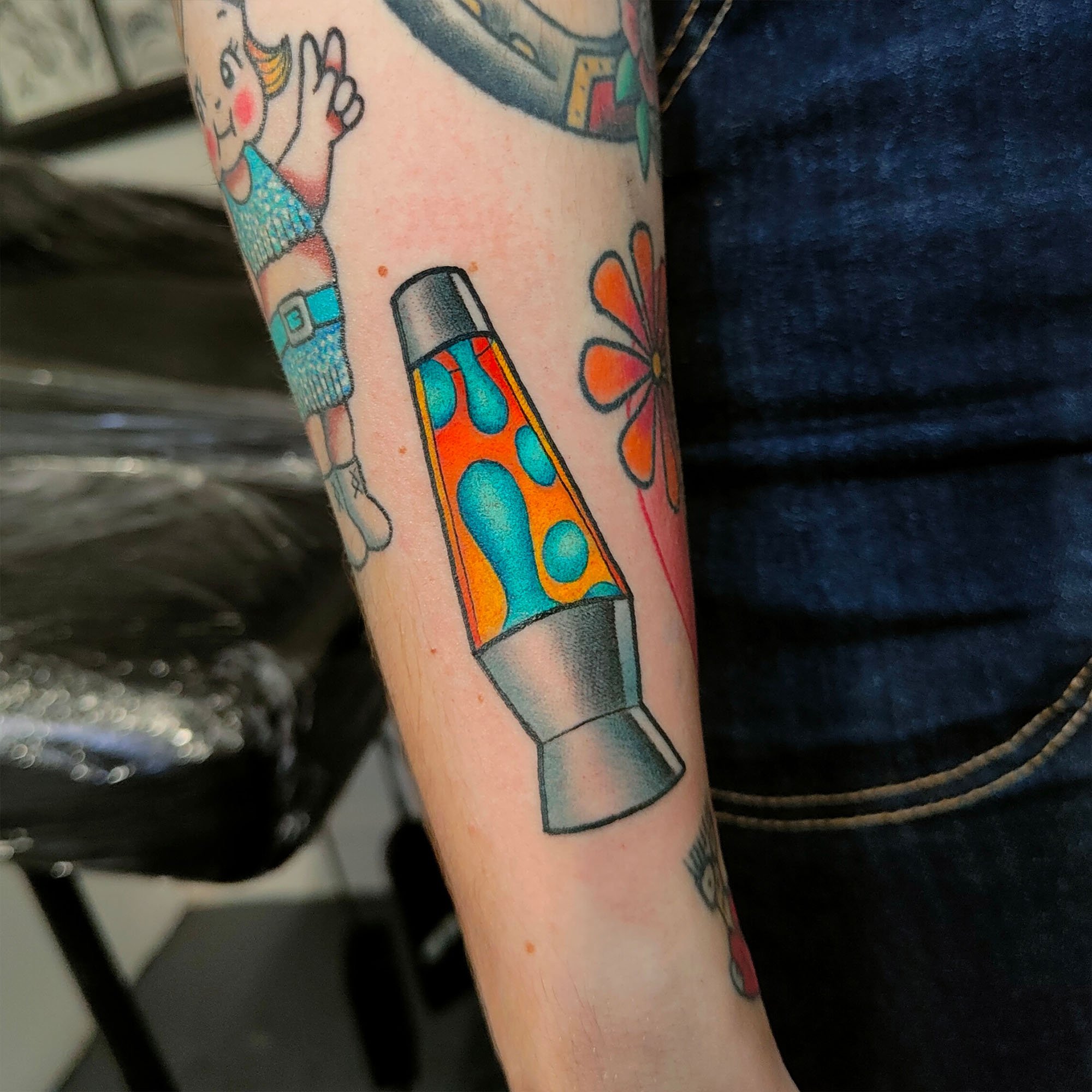 Tattoo of a colorful lava lamp on an arm with other tattoos including a cartoon character and flower design.