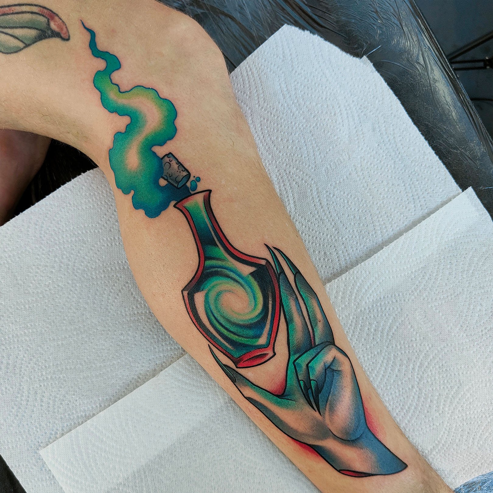 Colorful tattoo on a forearm featuring a stylized hand holding a swirling, iridescent potion bottle with green and blue smoke rising from it.