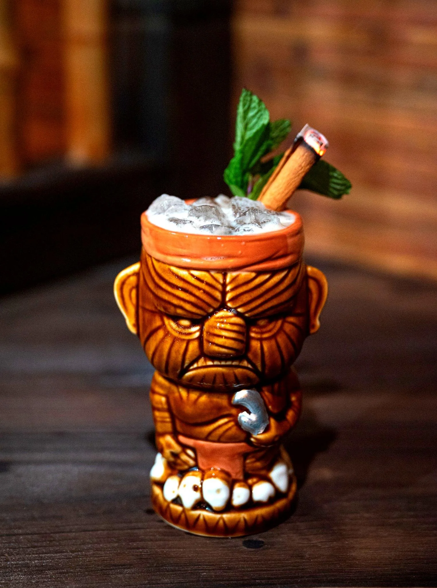 A tiki mug in the shape of a grumpy face with carved details, filled with ice, garnished with a cinnamon stick, mint leaves, and a metal straw, placed on a wooden surface.