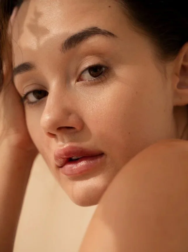 Close-up of a woman's face with makeup, looking over her shoulder with her hand resting on her head, light skin, dark hair, neutral background.