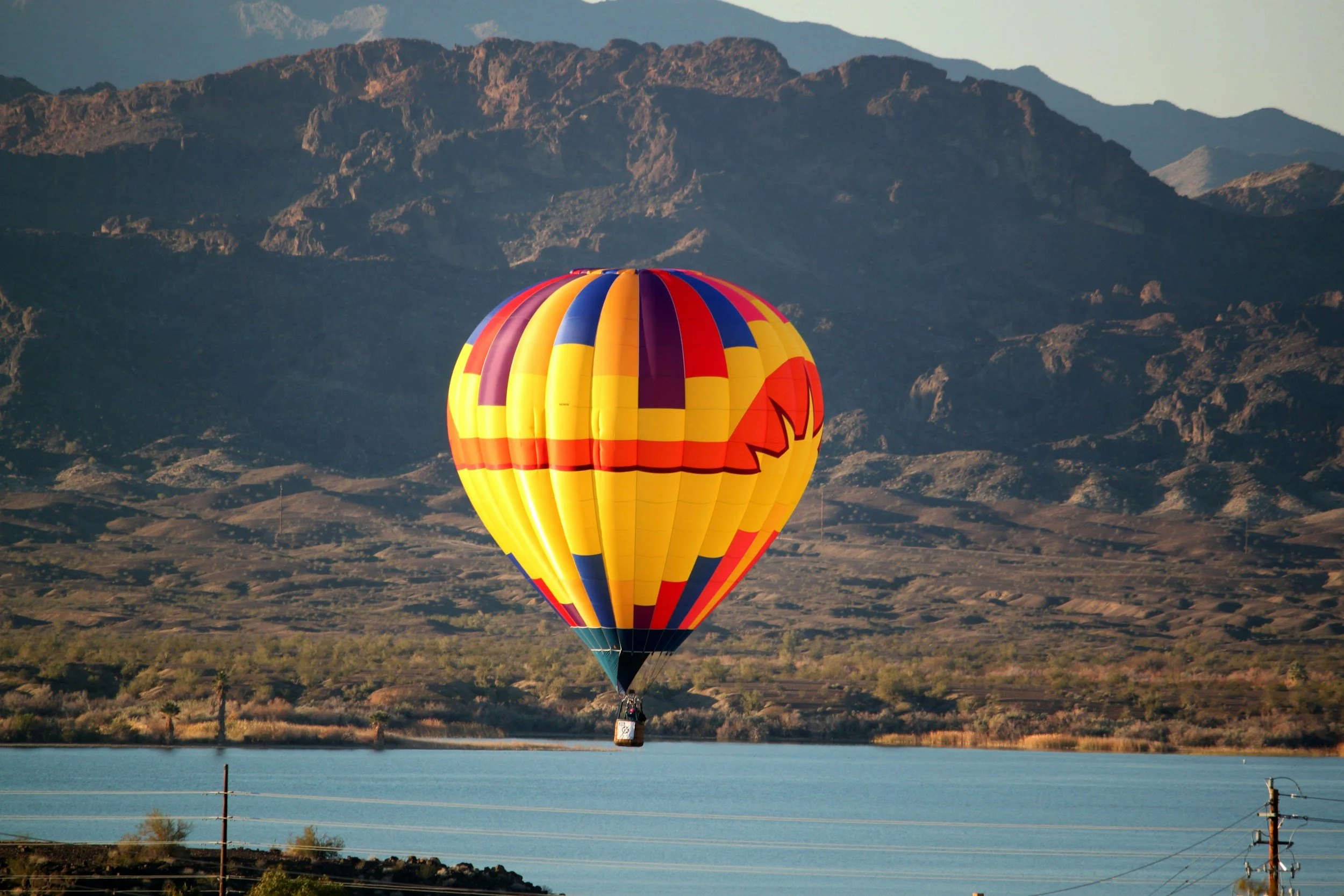 A colorful hot air balloon with yellow, red, purple, and orange panels floating above a body of water with mountains in the background.
