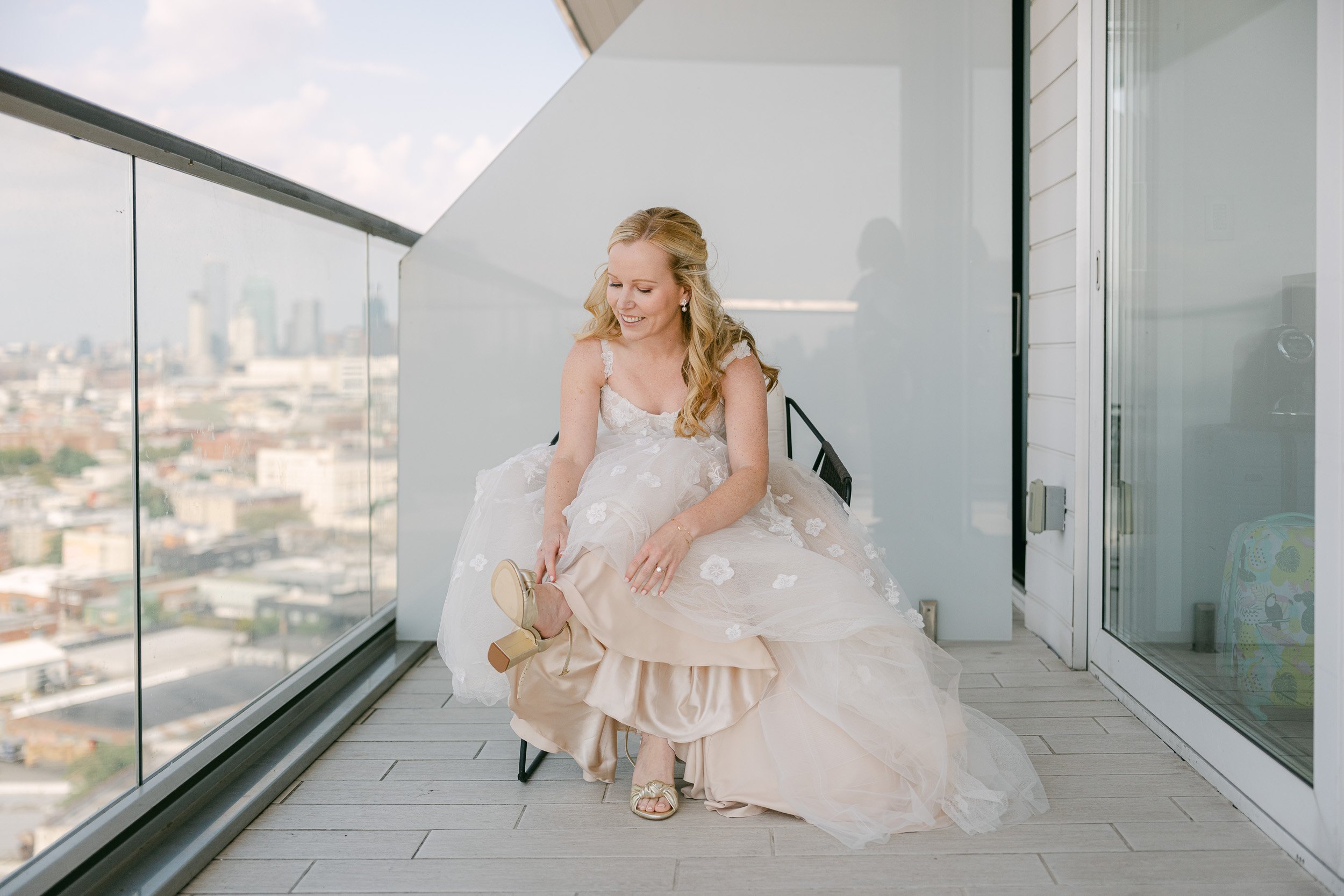 A bride in a wedding dress sitting on a chair on a balcony, adjusting her shoe. The balcony has glass railings with a cityscape view in the background.  This photo was taken in Williamsburg Brooklyn by Diane Stredicke - Hudson River Photographer