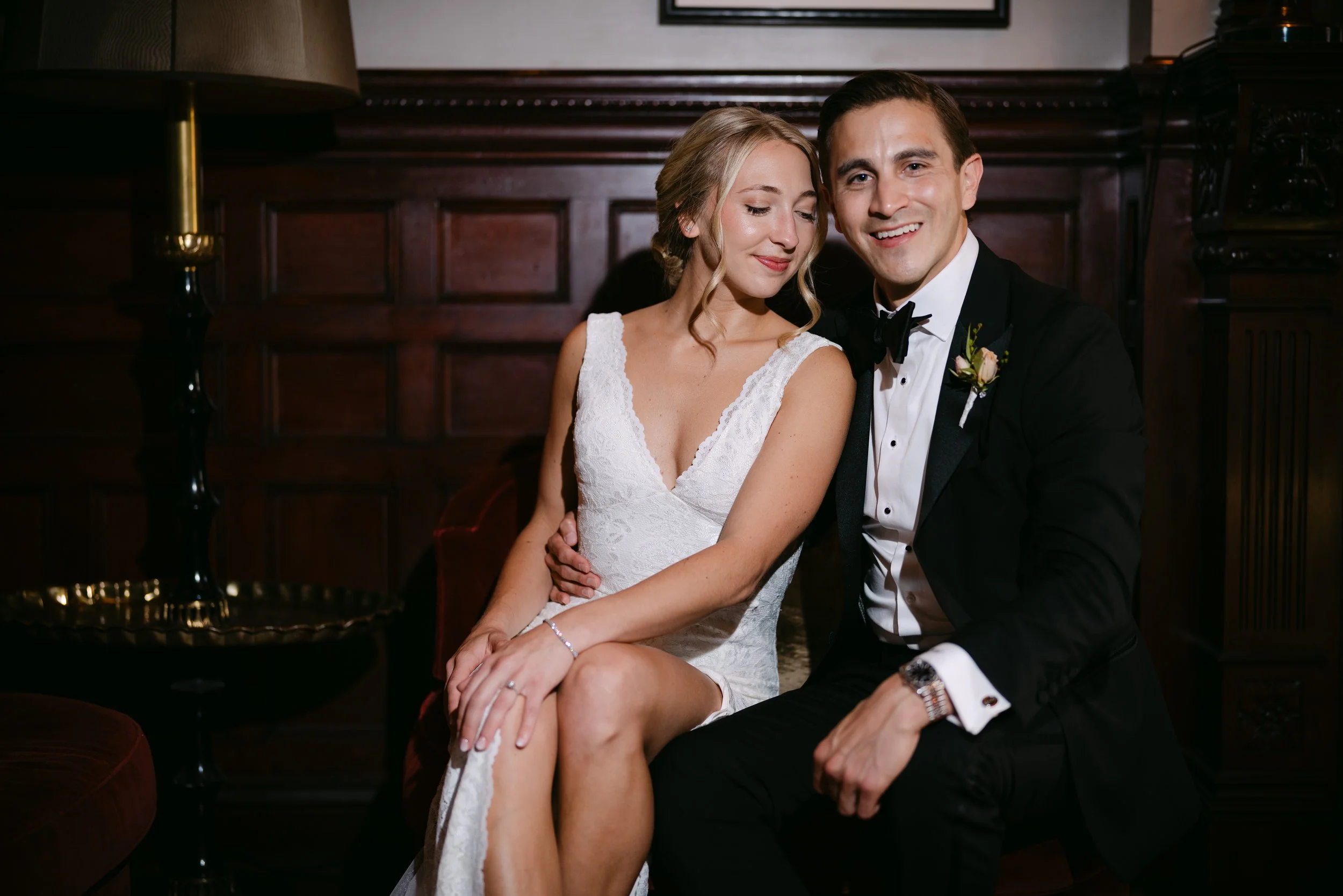 A bride and groom sitting close together in a dimly lit room. The bride is wearing a white lace dress, and the groom is in a black tuxedo with a bow tie. They are smiling with the groom looking at the camera and the bride with her eyes closed.