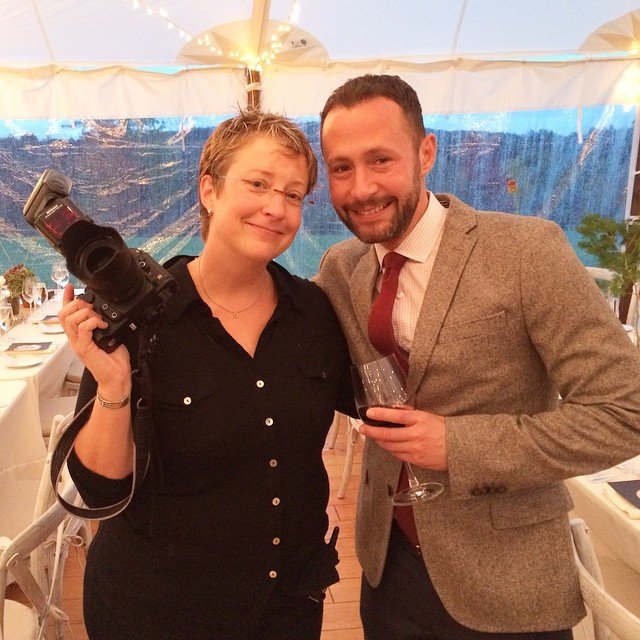 Award winning wedding photographer, Diane Stredicke, holding a camera and a man holding a glass of red wine at a social event, standing inside a tent with a scenic outdoor view in the background.