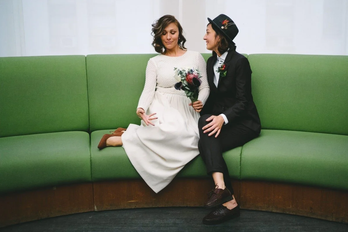 Two women sitting on a green couch, one in a white dress holding a bouquet, and the other in a black suit and hat, looking at each other.