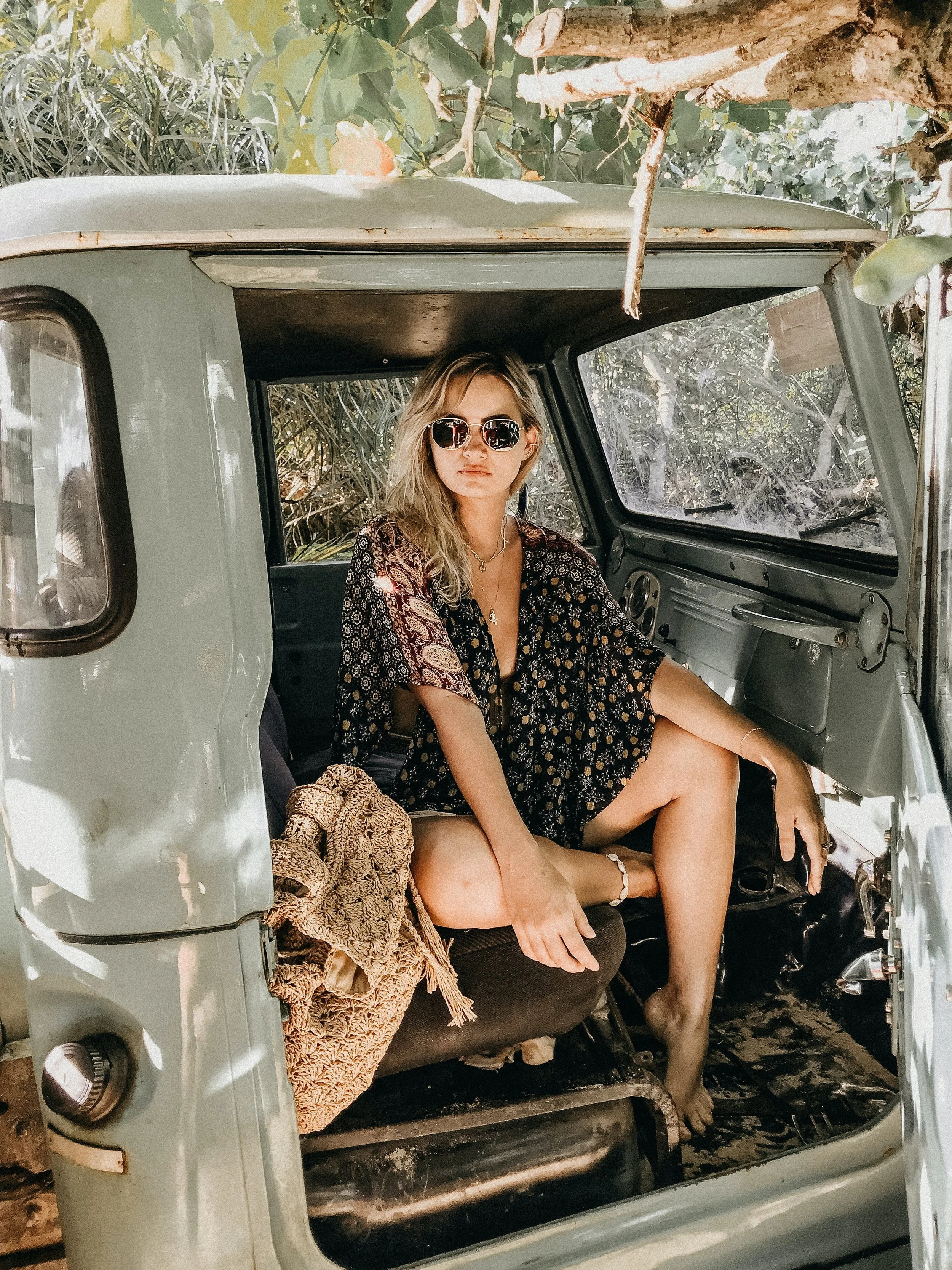 A woman with blonde hair and sunglasses sitting inside an old, weathered, green vehicle, with one leg crossed over the other, surrounded by leafy trees.