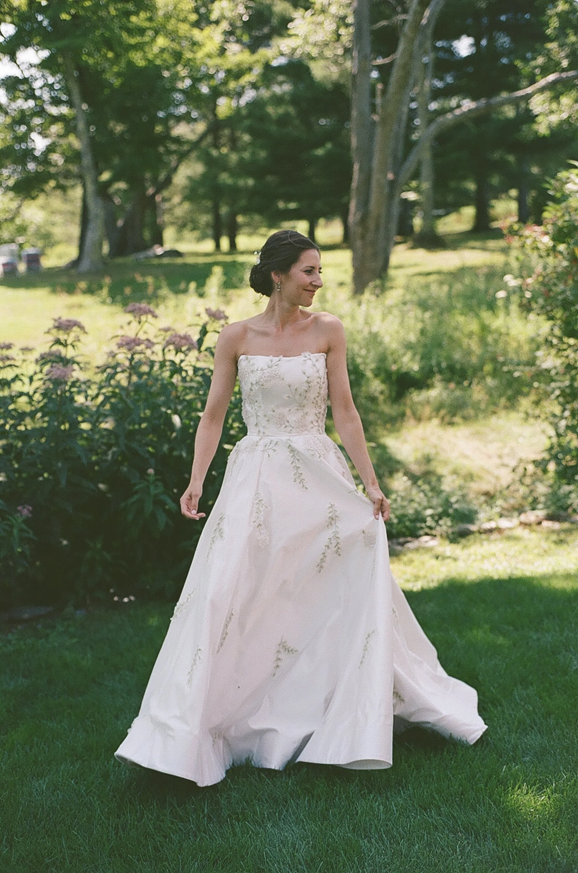 A woman in a white wedding dress standing outdoors in a garden with greenery and trees.