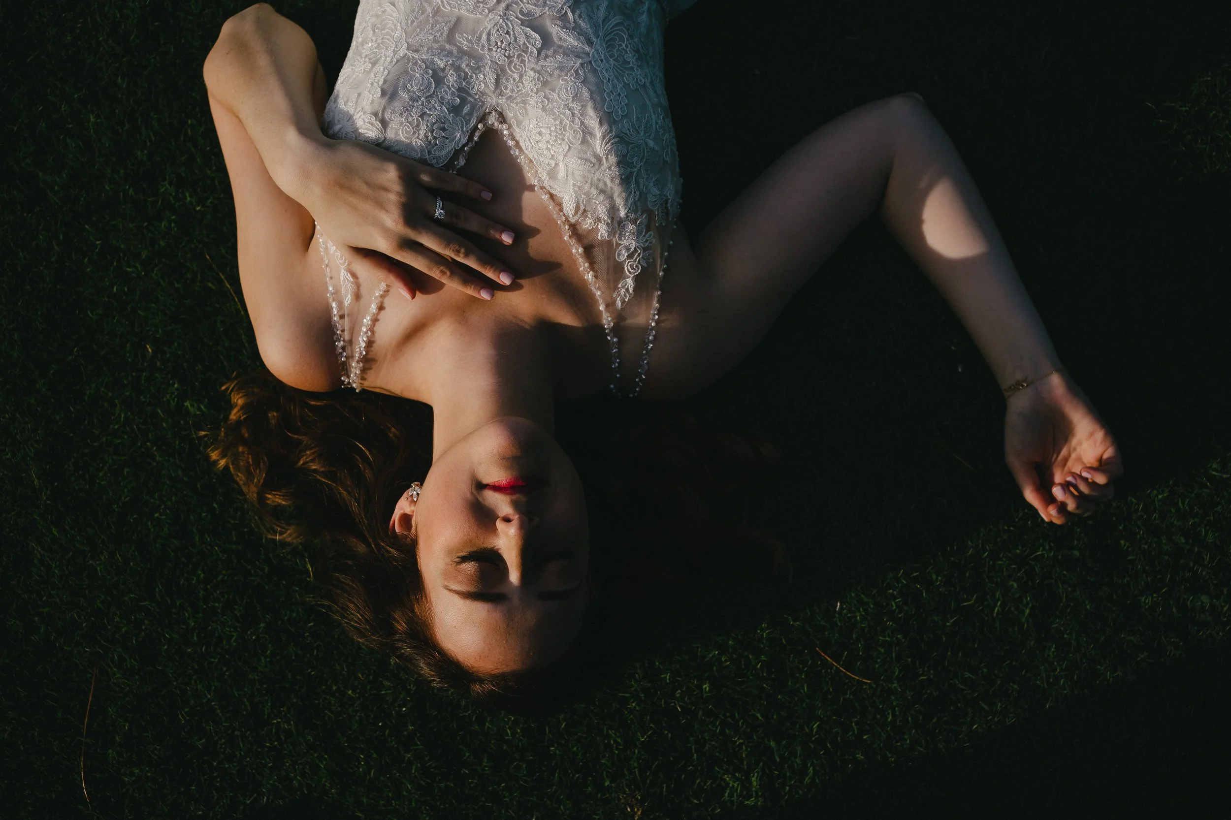 A woman with long, wavy hair lying on her back on grass, wearing a white lace dress, with her eyes closed and arm crossed over her chest.