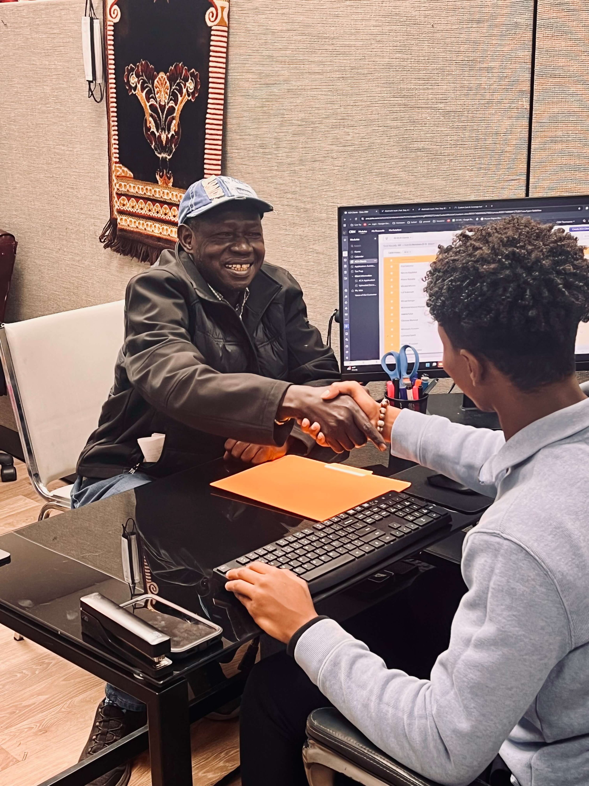 Two men shaking hands in an office, with a computer and office supplies on the desk.