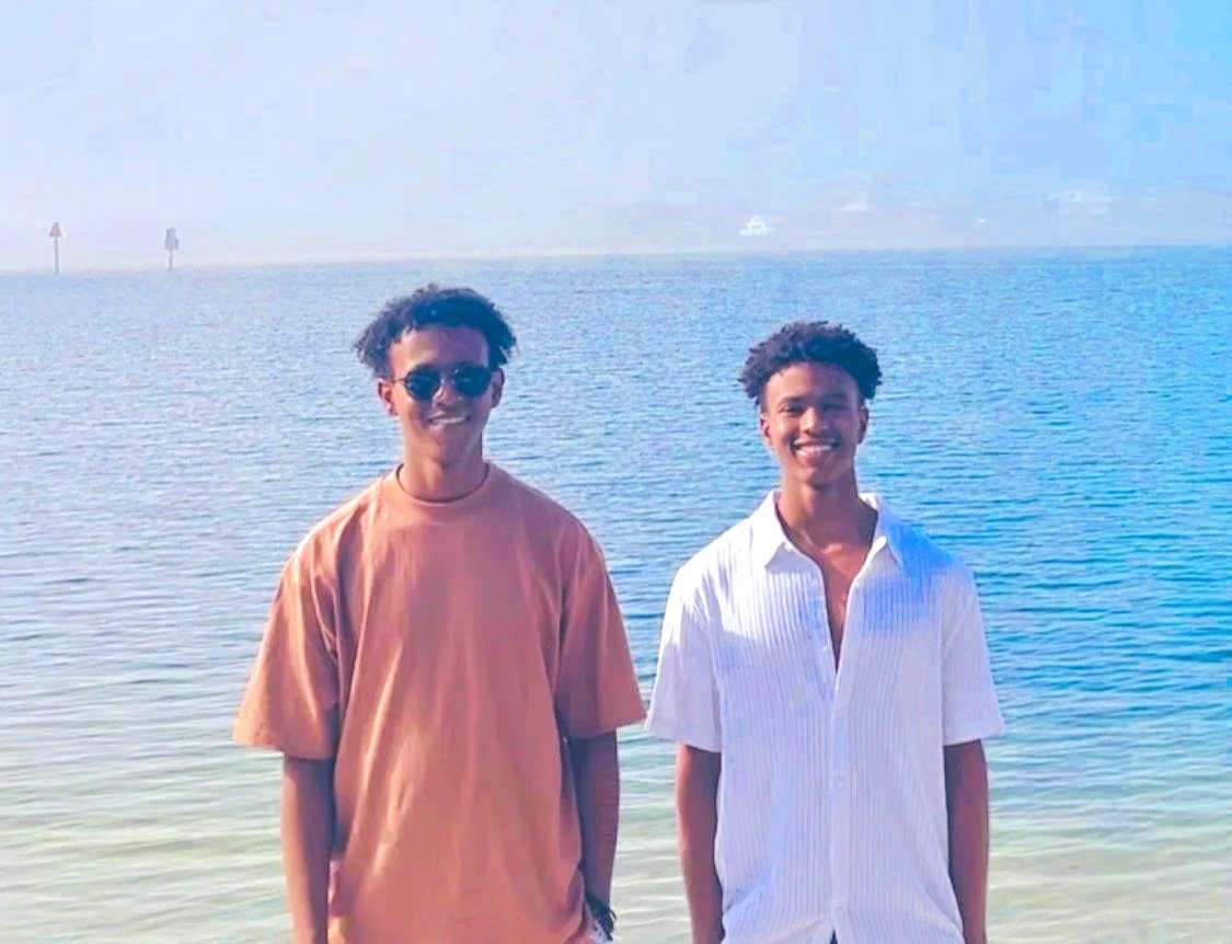 Two young men standing in front of a body of water, smiling at the camera. One is wearing sunglasses and a tan t-shirt, the other is in a white button-up shirt. The water appears calm and there are some boats in the distance.
