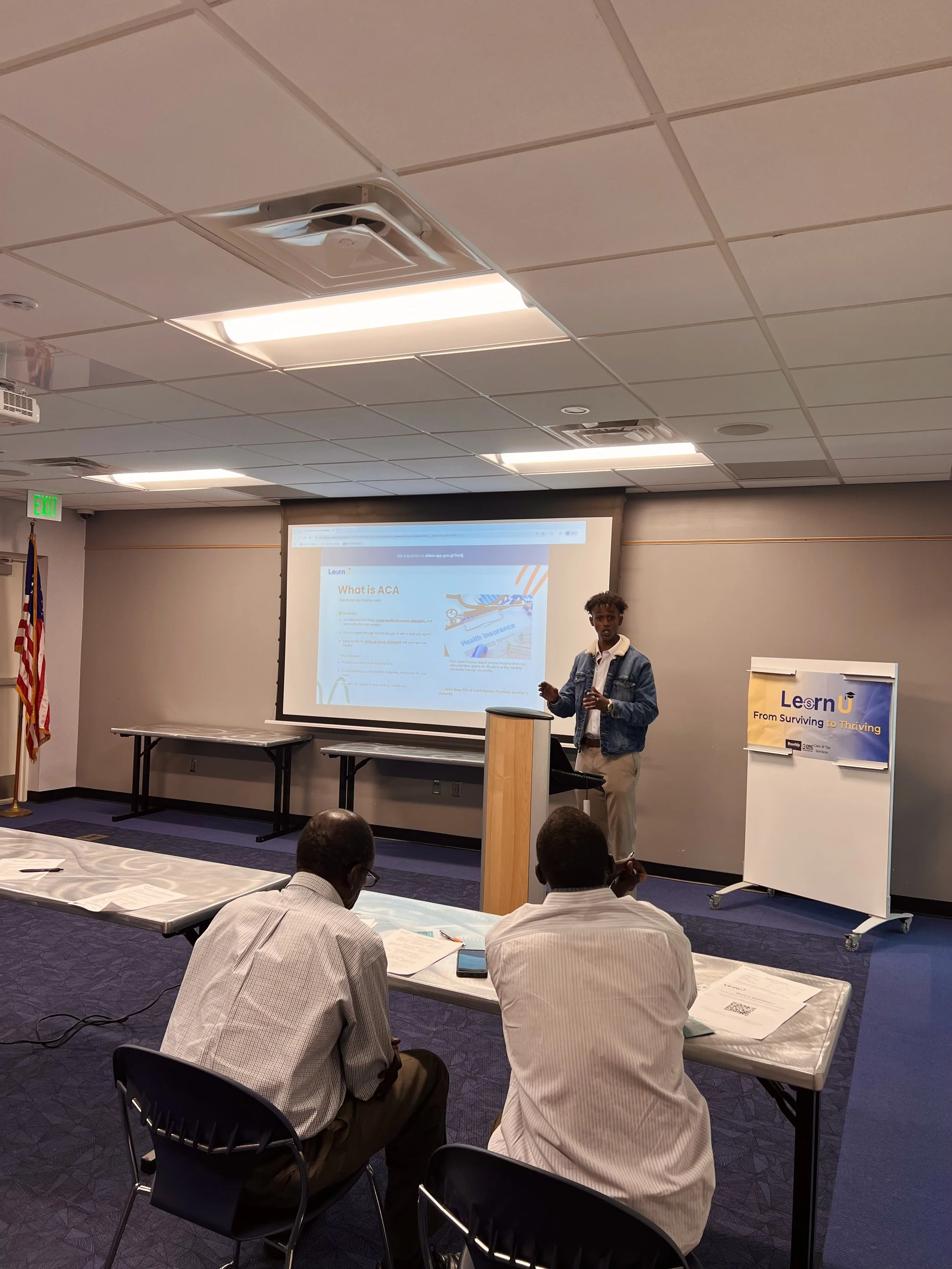 A man giving a presentation in a conference room with a slide titled 'What is ACA' displayed on a screen. Two people are seated at a table in the foreground, facing the speaker.