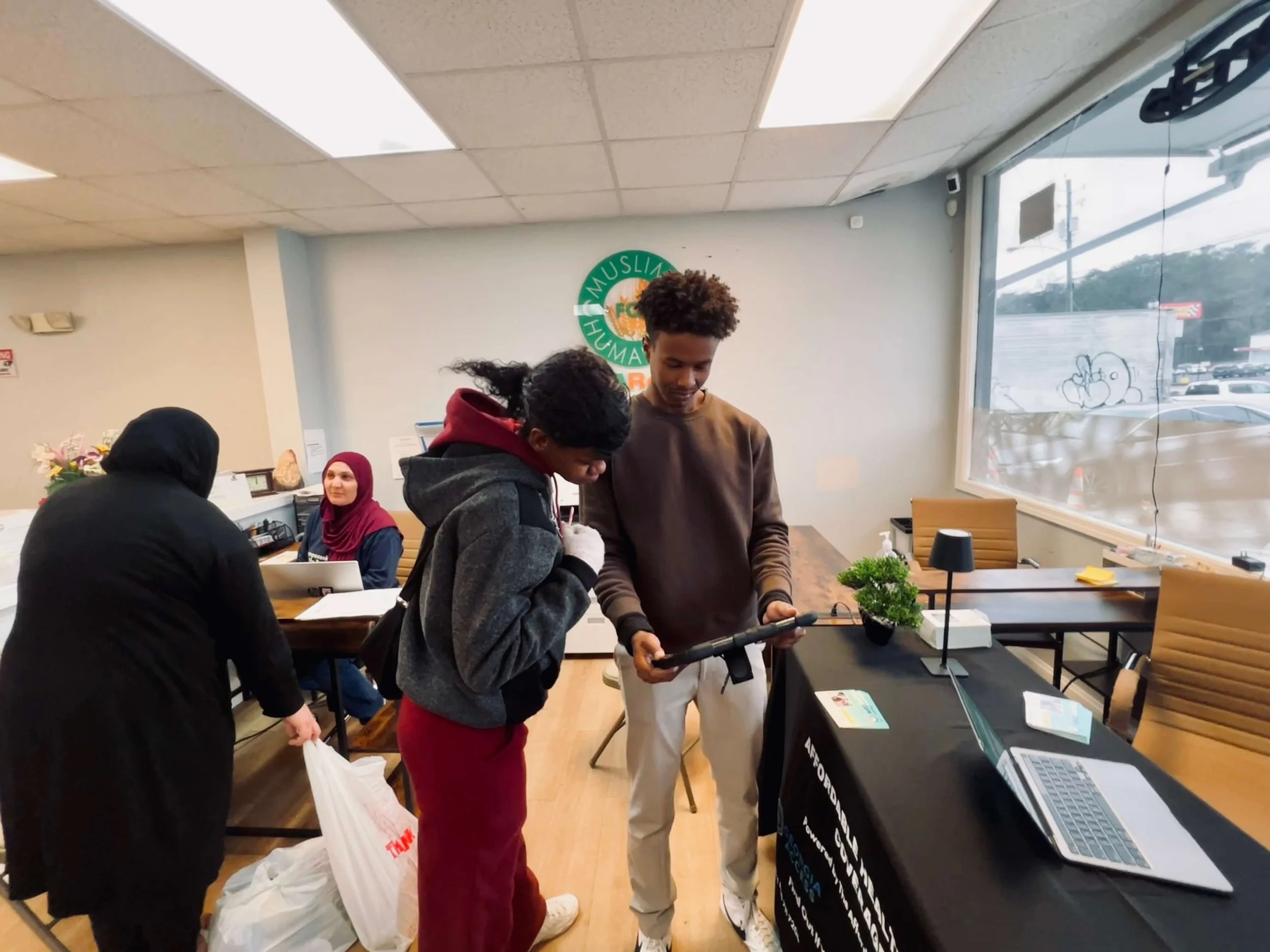Two young men are engaged in a conversation inside a community center. One is holding a tablet and the other is looking at it. There is a woman sitting at a desk in the background and another woman wearing a headscarf nearby. The room has large windows, chairs, and a sign on the wall that reads 'Muslim Human', with the full sign likely being 'Muslim Humanitarian'.