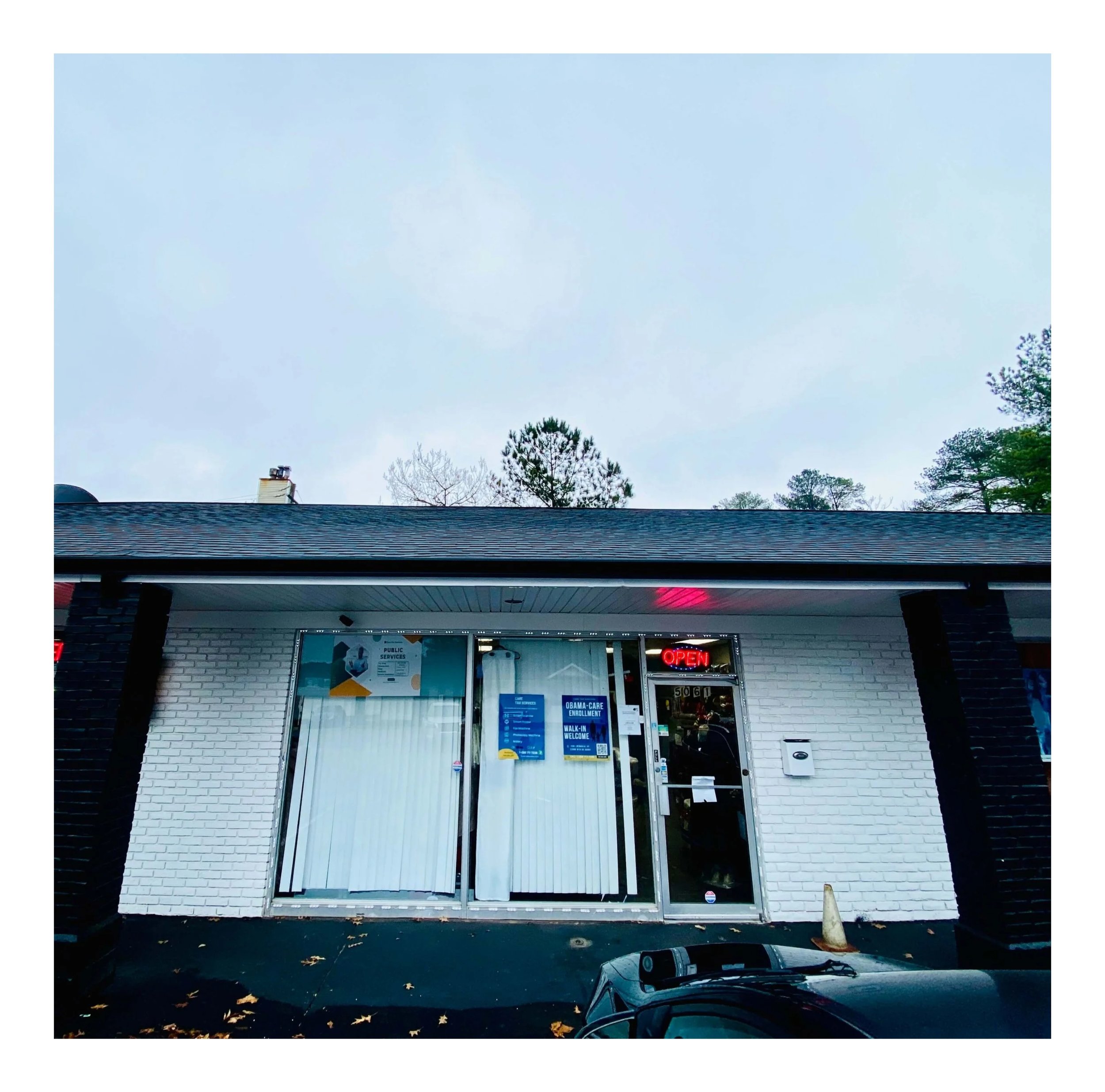 Exterior view of a small office building with white brick walls, blue signs, glass doors with vertical blinds, and an illuminated red 'OPEN' sign. A traffic cone is placed outside near the entrance, and a car is partially visible in the foreground. The sky is overcast with trees in the background.