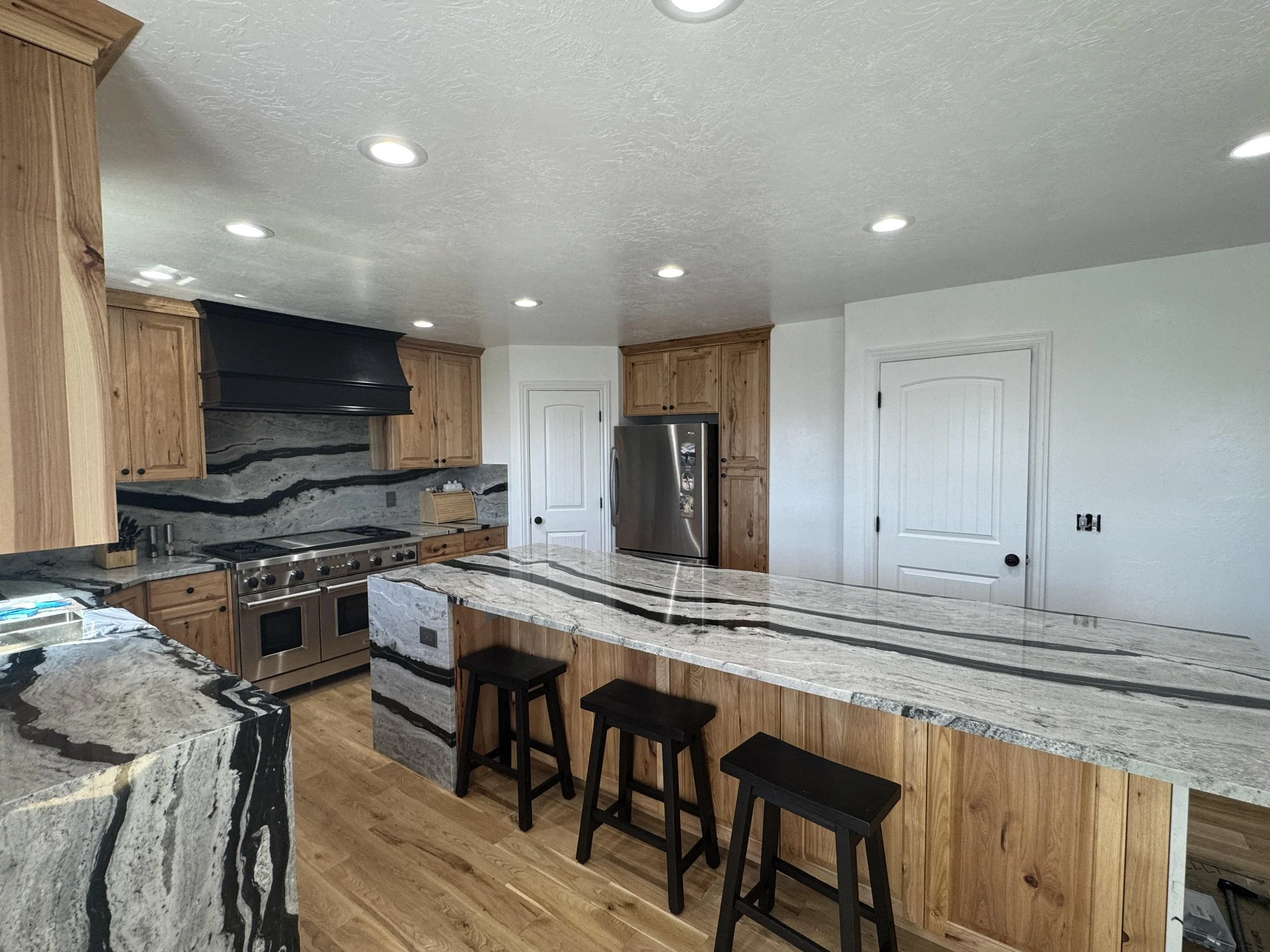 A modern kitchen with Hickory cabinets, a marble island, stainless steel appliances, including a refrigerator, and a black range hood. The ceiling has several recessed lights, and there are two black barstools at the island.