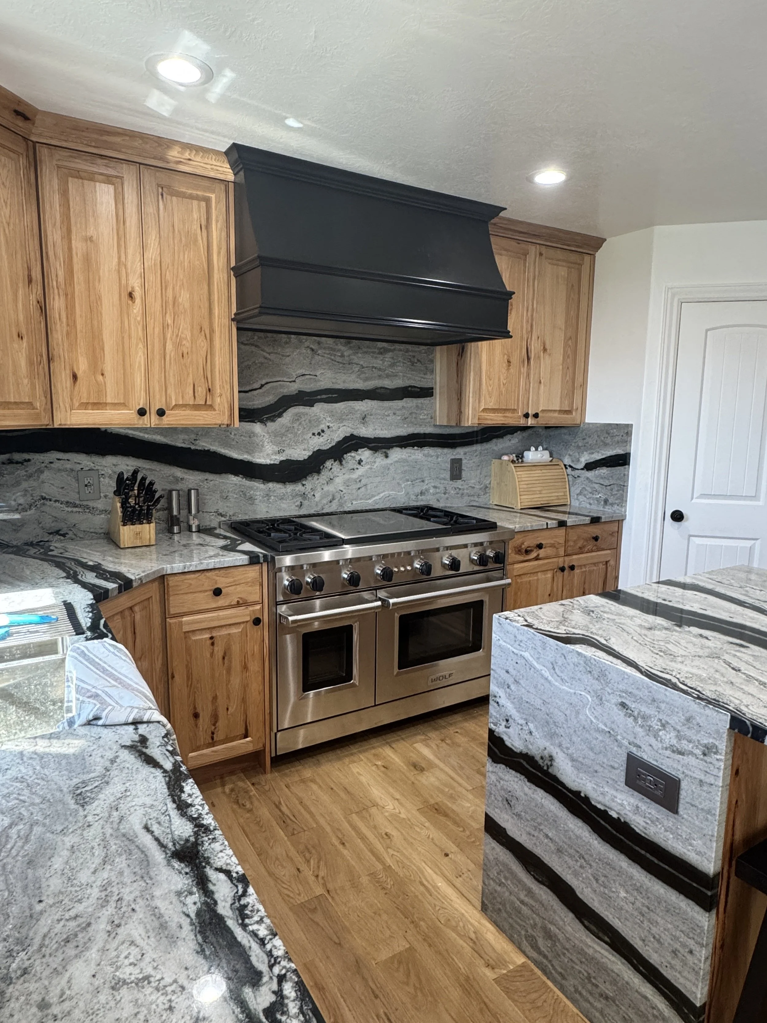 This is a hickory Kitchen, a paneled granite countertop and backsplash with black, gray, and white swirls, a stainless steel stove and oven, a knife block on the counter, and a bread box. There are reccessed ceiling lights and hardwood flooring.