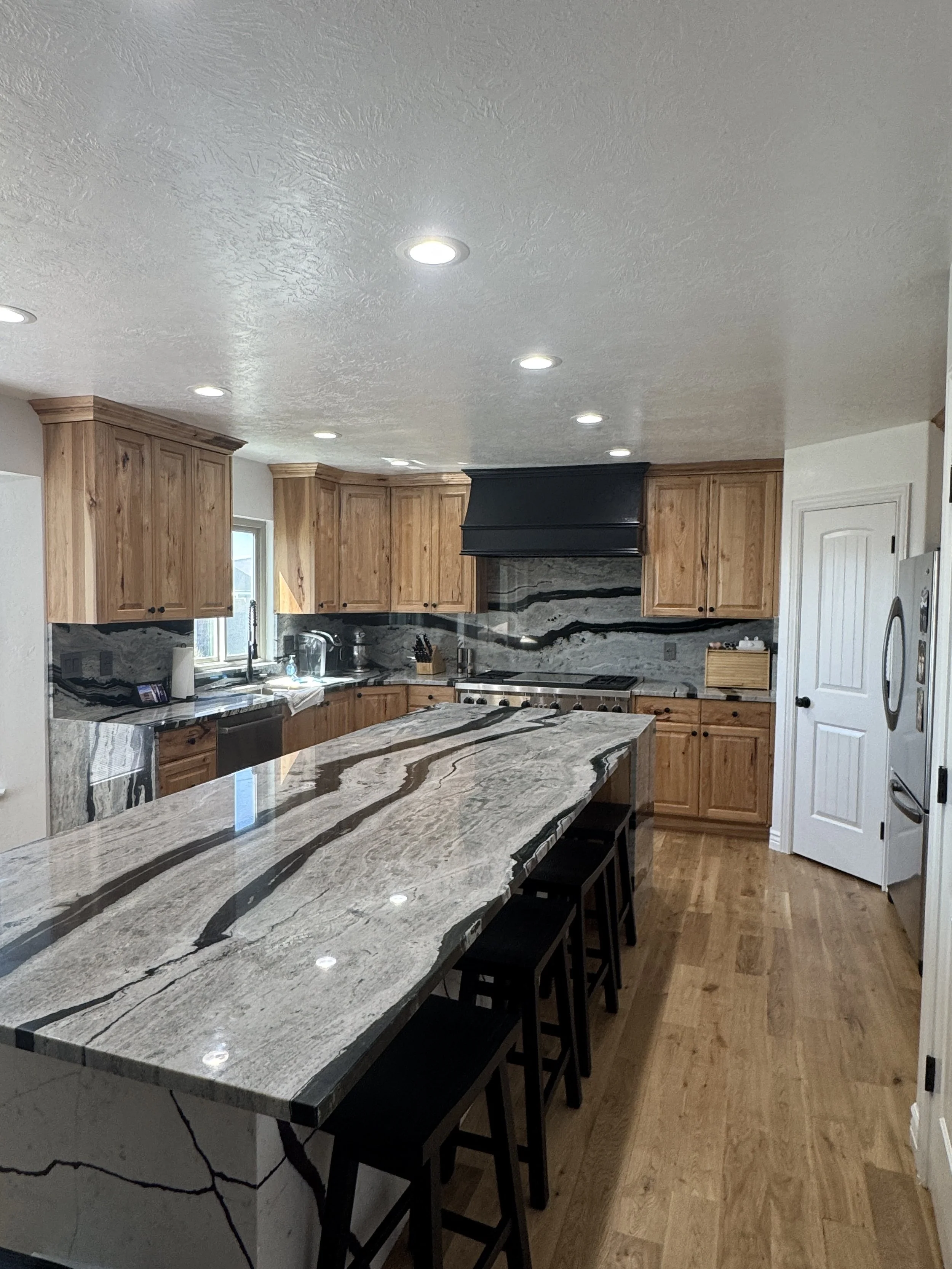 Kitchen with Hickory wooden cabinets, a marble countertop island, a black range hood, stainless steel appliances, and hardwood floors.