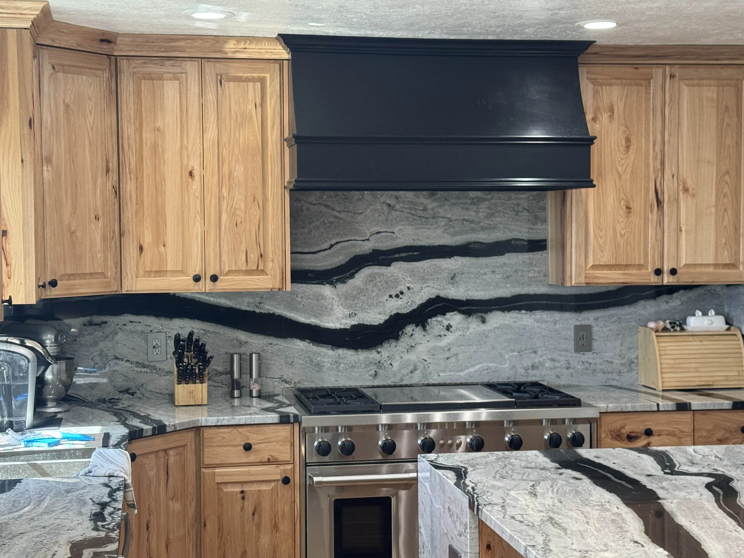 Custom Kitchen with Hickory cabinets, granite countertops, a stainless steel stove, and a custom black range hood. There are utensils and a toaster on the counter, and a marble backsplash with black, white, and gray patterns.