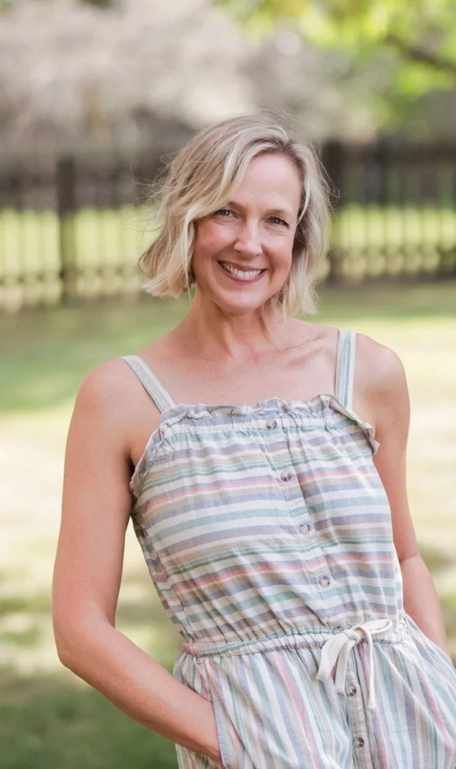 Smiling woman in a striped summer dress outdoors with a blurred wooden fence and trees in the background.