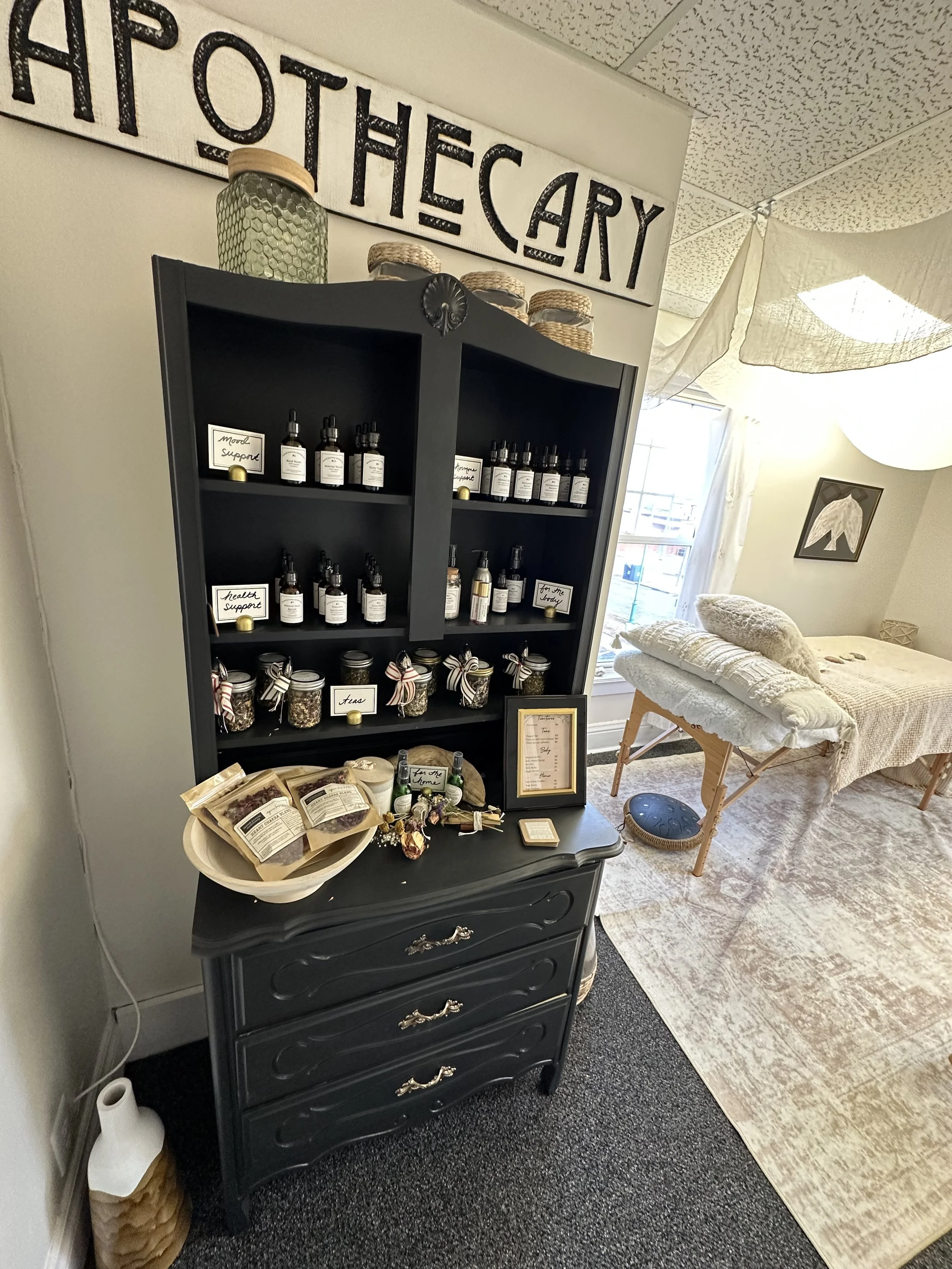 Four amber glass dropper bottles labeled from Sacred Journey Apothecary, with remedies for sleep, mood boost, anxiety relief, and natural pain relief, set on a wooden surface with a beaded necklace in front, surrounded by potted plants in the background.