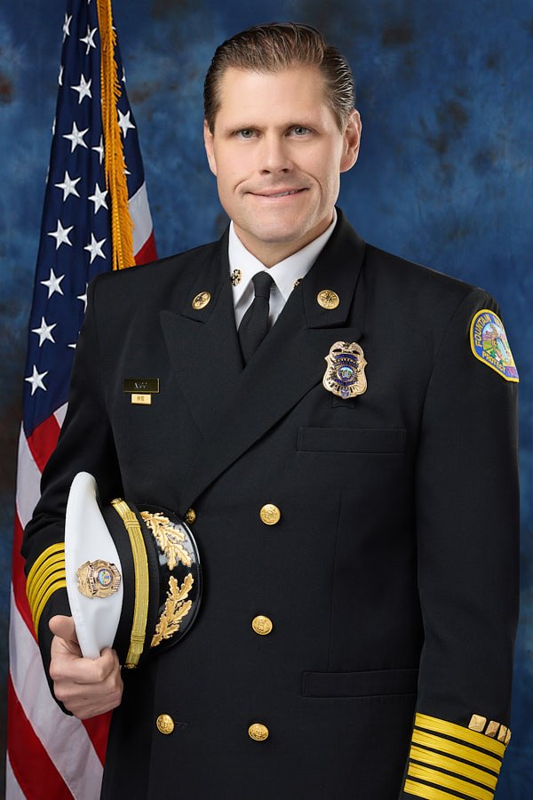 A fire chief in uniform standing in front of an American flag with a blue background, holding a white fire officer cap with gold embellishments.