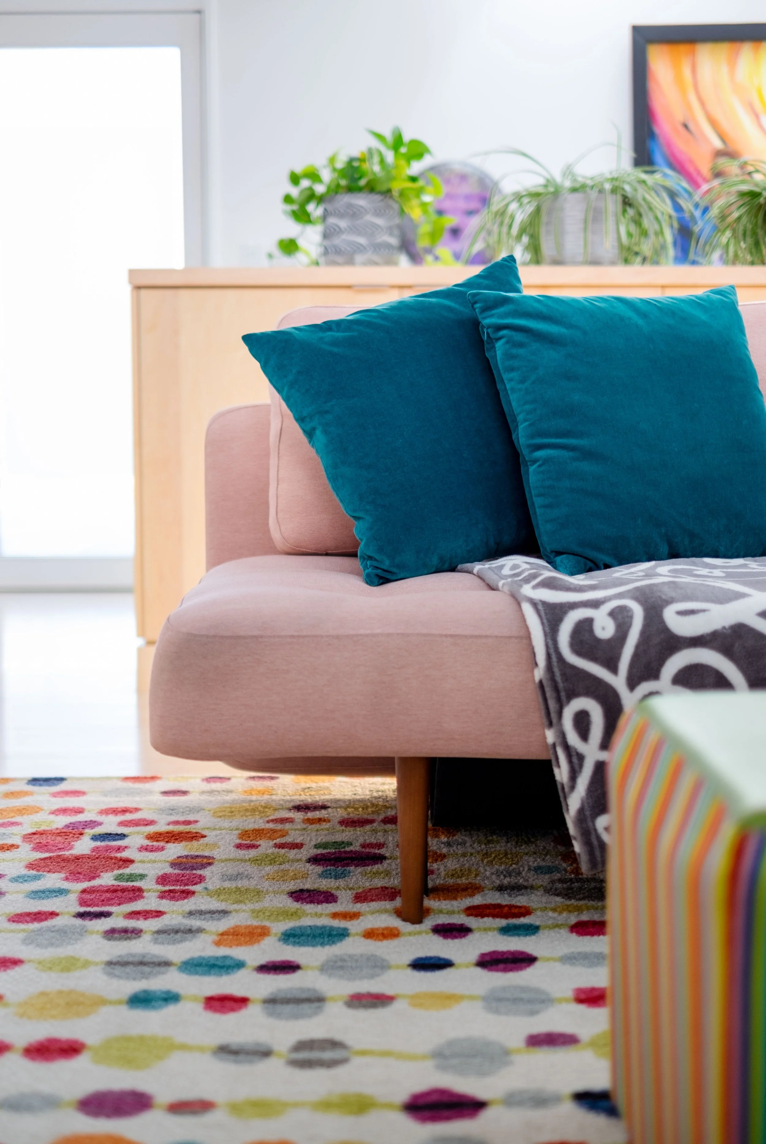 Modern living room with a pink sofa and teal pillows, a colorful polka dot rug, and potted plants on a wooden cabinet.