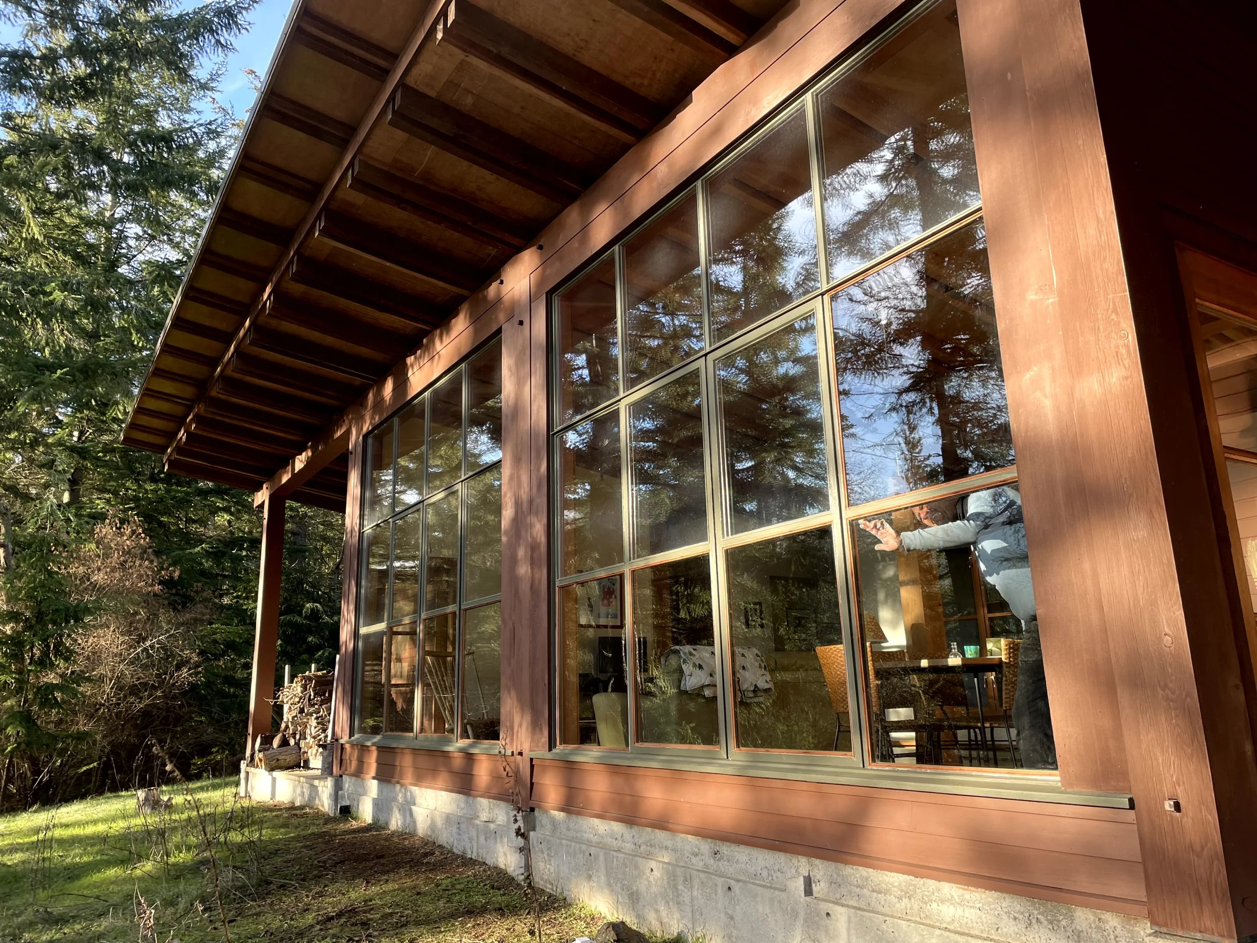 A person inside a modern wooden house, seen through large glass windows, reaching out with both hands. The house is surrounded by trees and greenery.