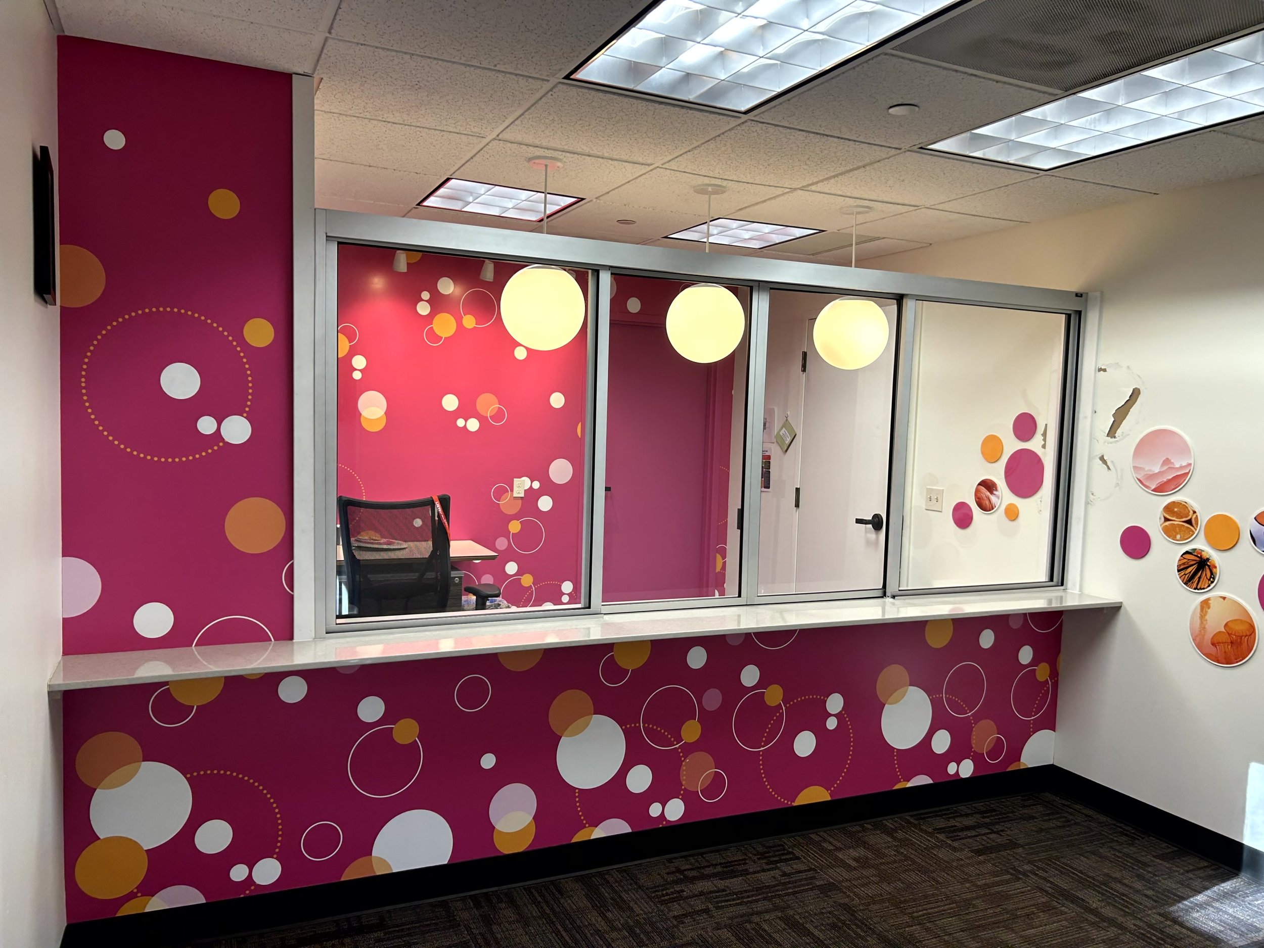 Office reception window with pink and white bubble patterned walls and a counter. Behind the window is a chair and desk, with pink walls decorated with circular designs. To the right, a white wall displays mounted circles with various images, and the