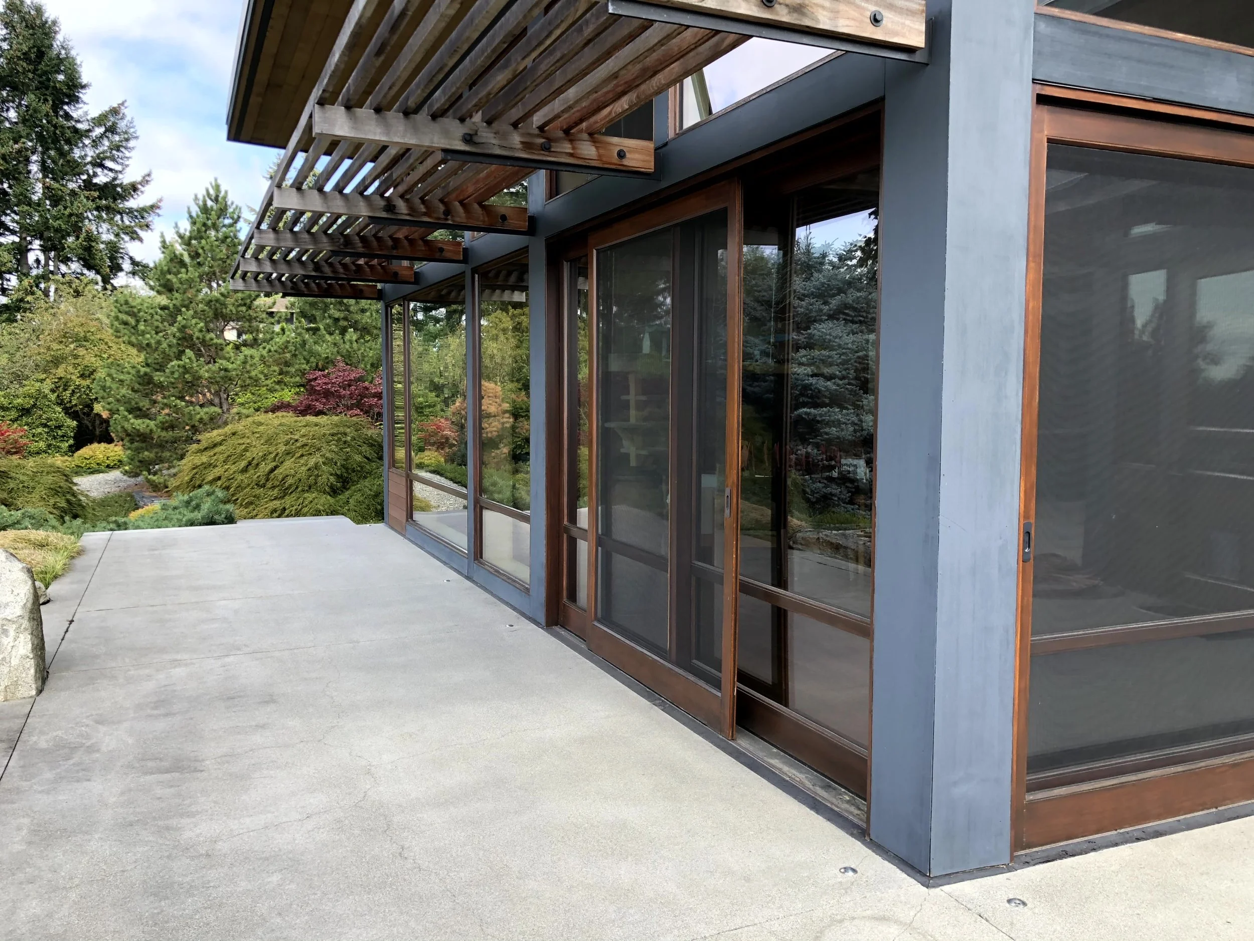 View of a modern house exterior with large glass sliding doors, a concrete patio, and a Japanese garden with lush trees and shrubs.