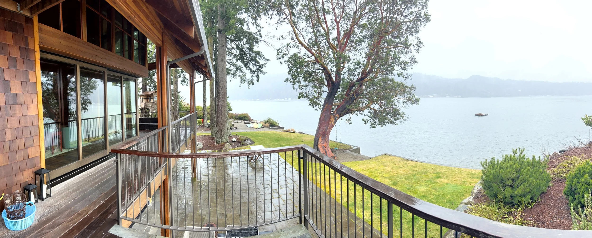 View from a wooden deck overlooking a lake with a tree, boat, and mountains in the background.