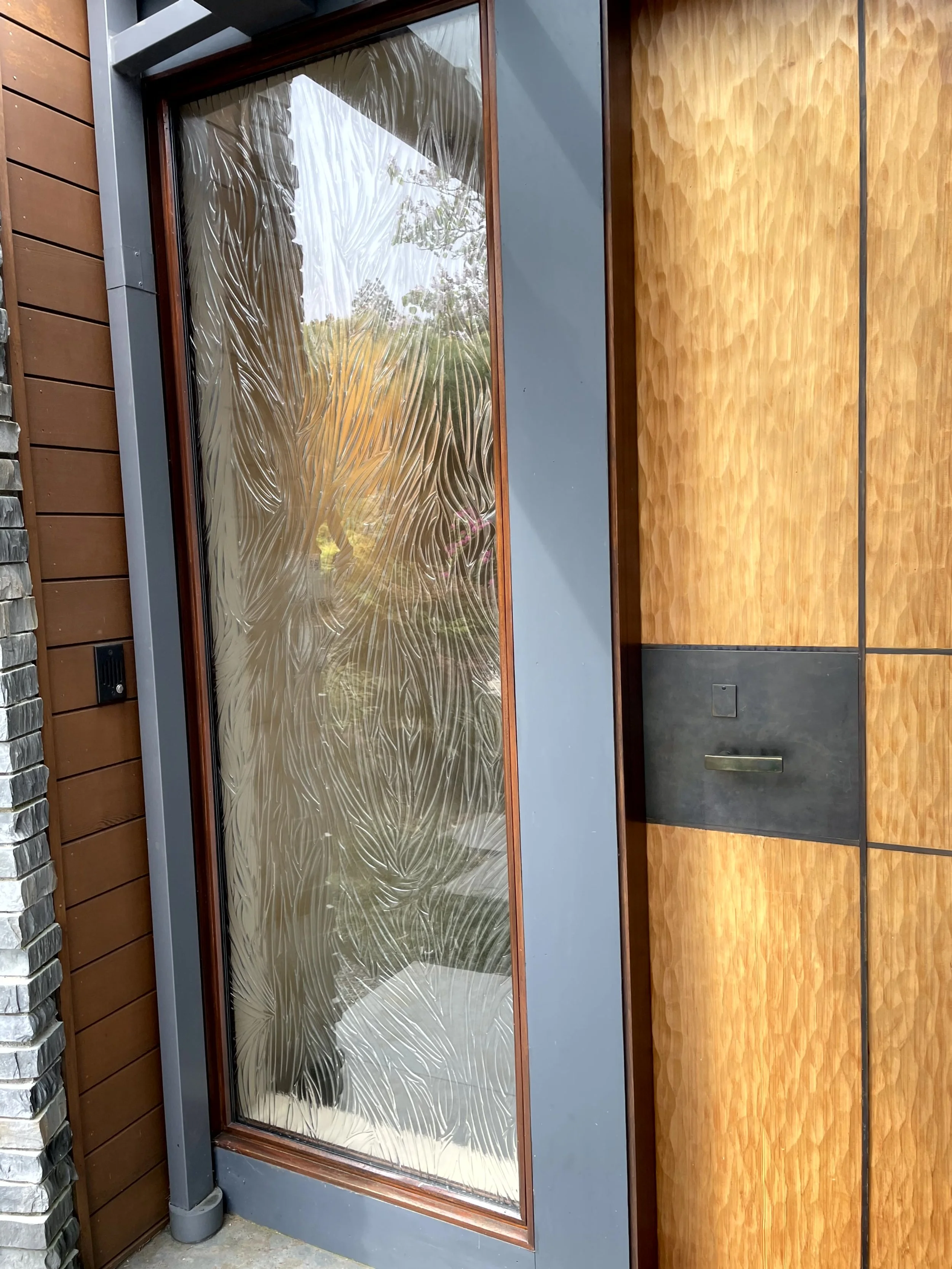 Decorative glass panel door with an etched leaf pattern, set in a metal frame, next to wooden paneling on a building exterior.