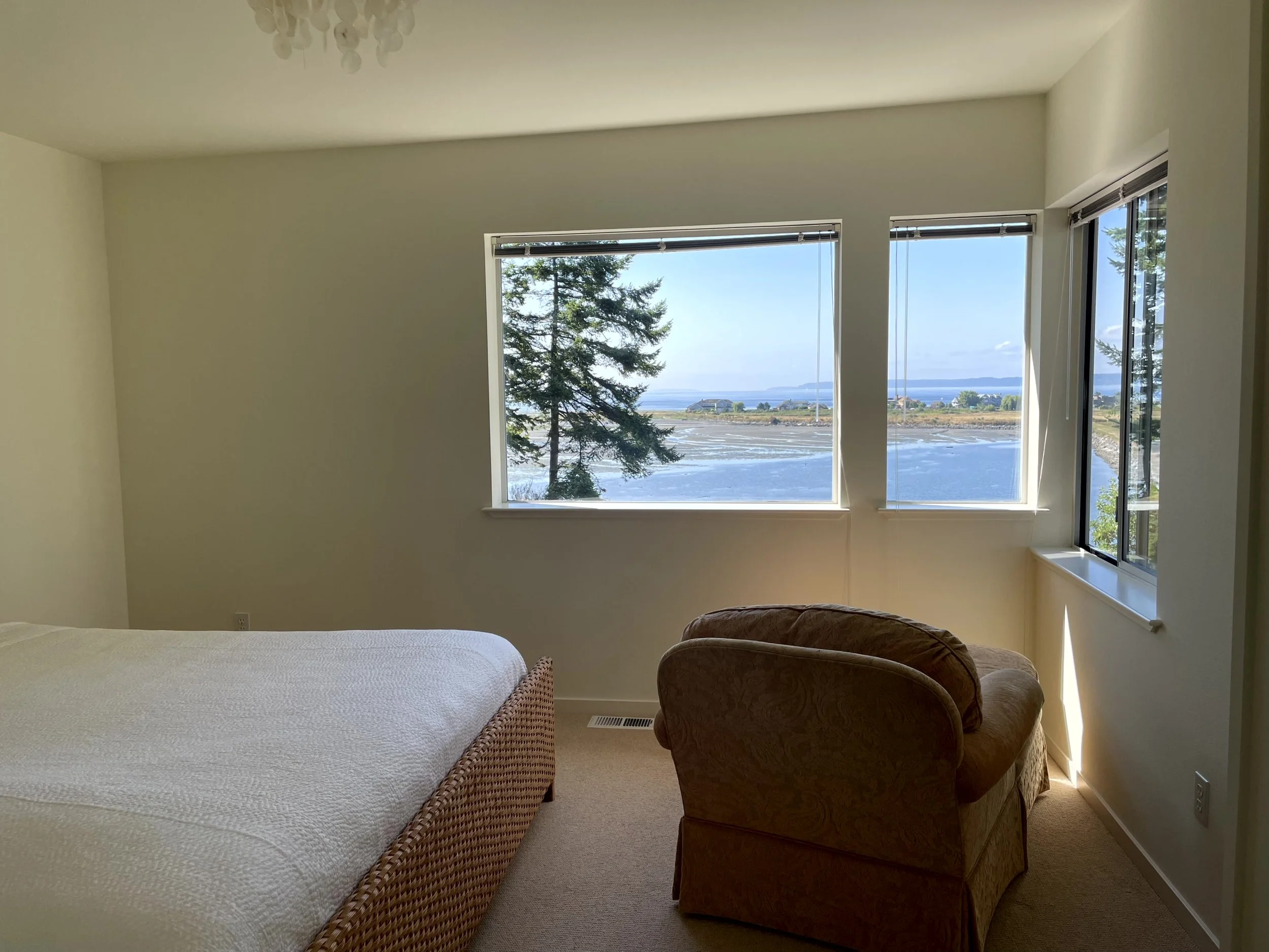 A bedroom with a large window showing a water view and trees outside; a bed with a wicker frame and white bedding, and a patterned armchair near the window.