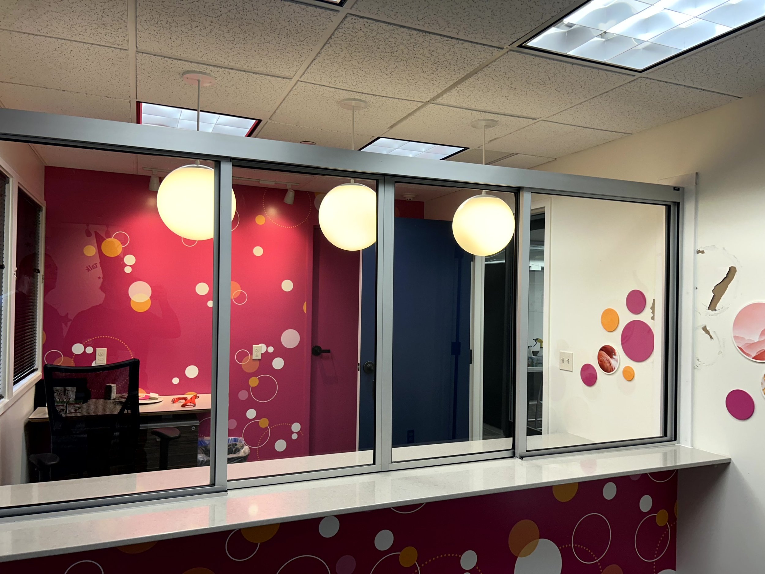 Interior of an office cubicle with pink and blue walls decorated with circles, three hanging ceiling lights, and a white countertop in the foreground.