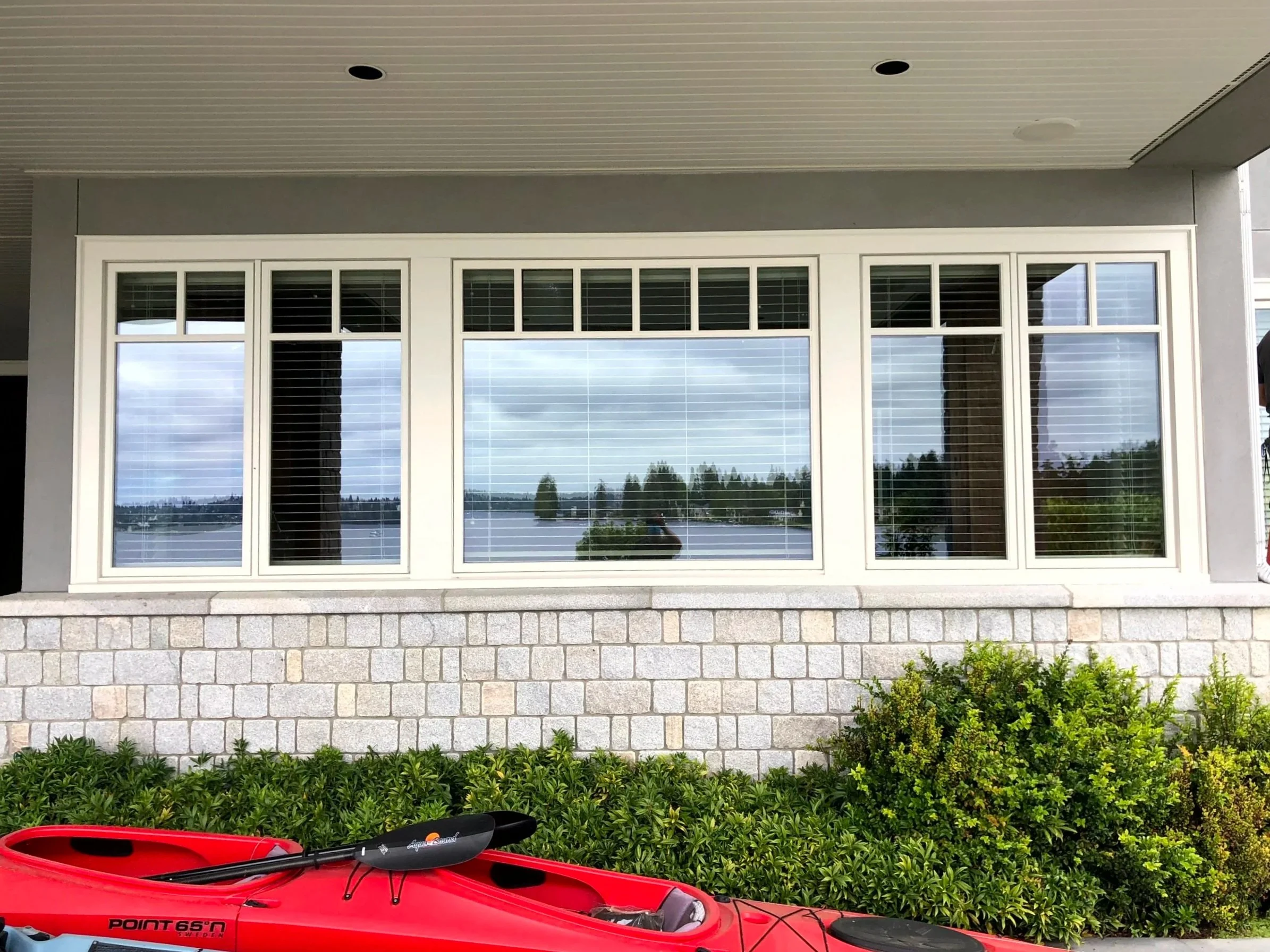 A large window with multiple panes and white framing on a house exterior, reflecting a cloudy sky and trees, with a red kayak and paddle on green bushes below.