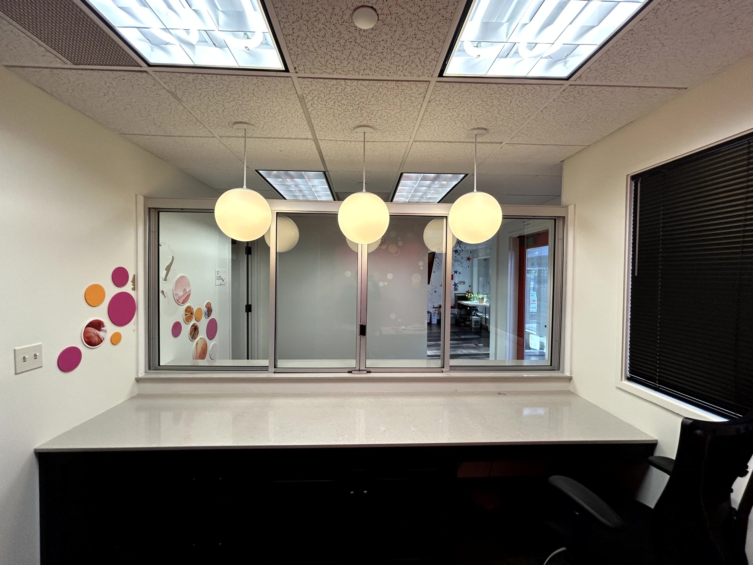 Empty office or conference room with a long white table, black office chair, black blinds, and a window wall with frosted glass and colorful wall decorations, illuminated by three hanging round lights.