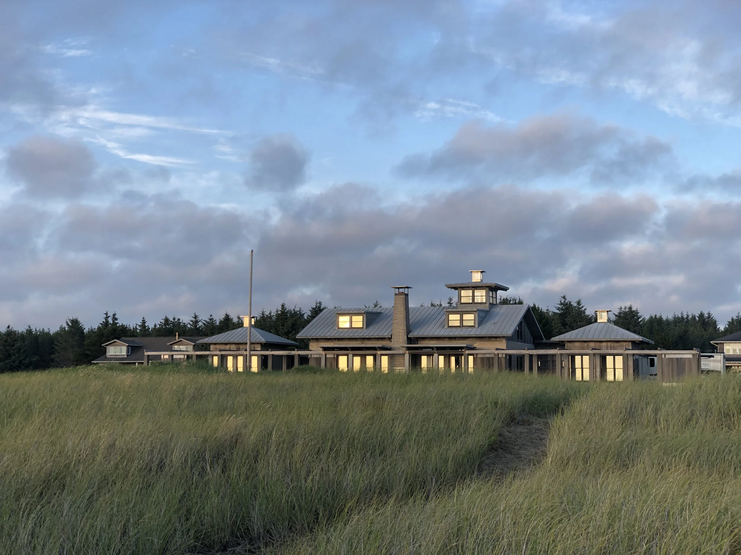 A large house with multiple roofs and a chimney, surrounded by tall grass, under a partly cloudy sky.