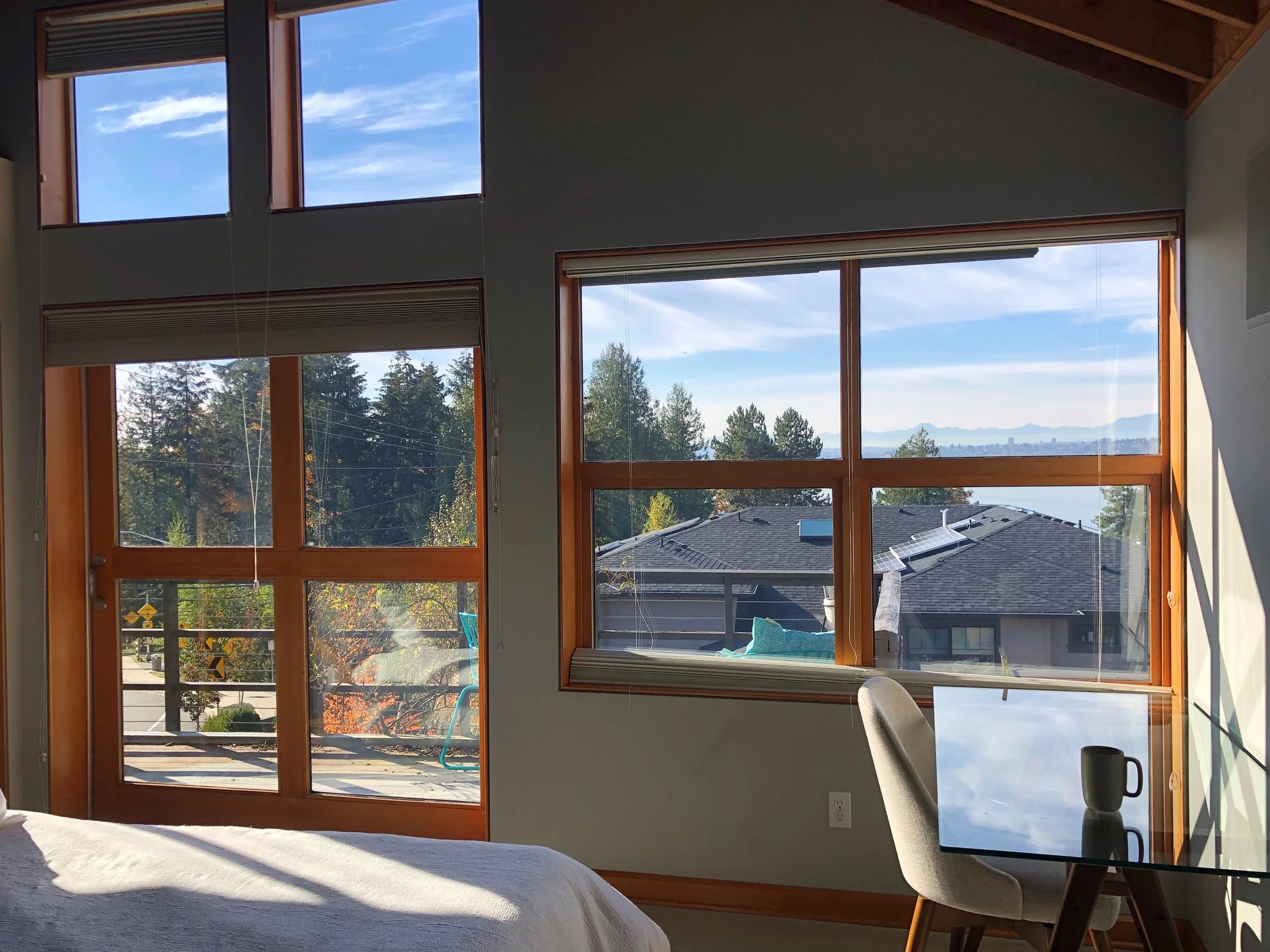 Interior view of a bedroom with large windows showing trees, rooftops, and a scenic view of the ocean or lake in the distance. The room includes part of a bed, a small glass table with a coffee mug, and a chair.