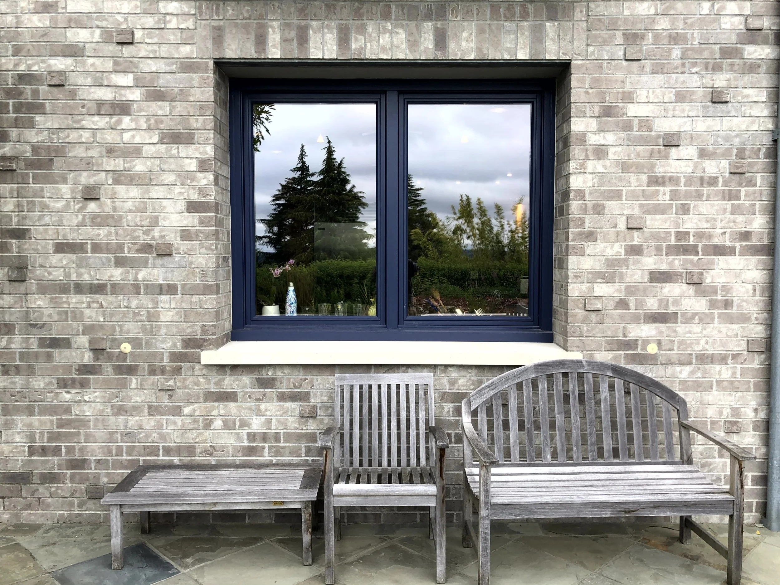 Outside view of a brick house with a large double window, flanked by two wooden benches and a small table on a stone patio.