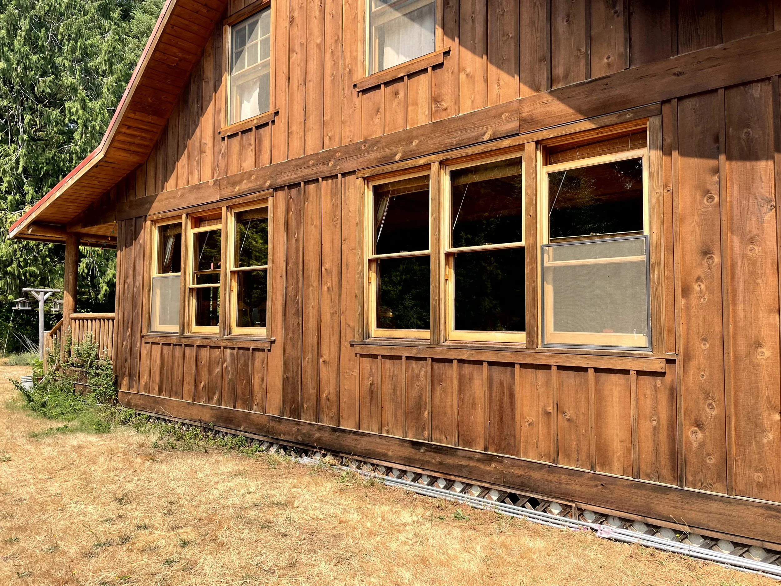 Wooden house with multiple windows and a sloped roof, surrounded by trees and dry grass.